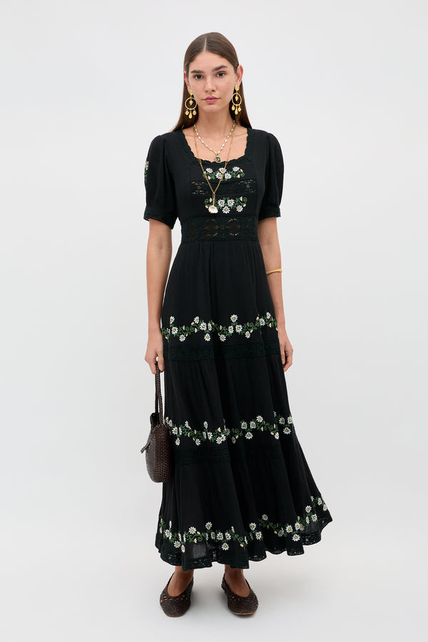 A woman stands against a plain light background wearing the Philippa - Lilia Embroidery Black dress with white floral details, gold statement earrings, layered necklaces, brown shoes, and a brown woven handbag.