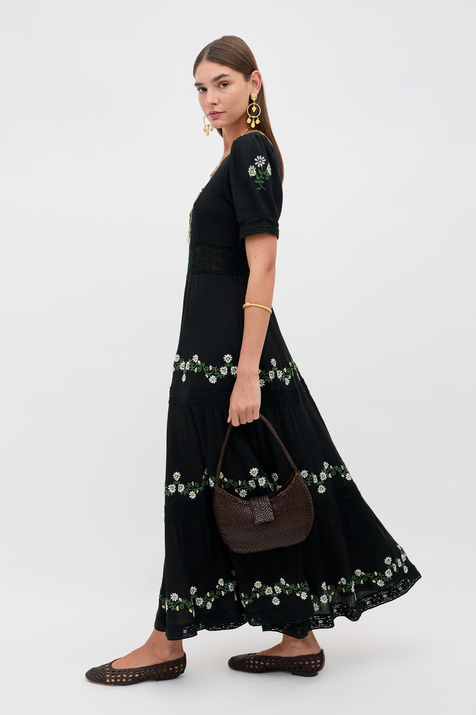 A woman wears the Philippa - Lilia Embroidery Black dress, styled with a brown woven handbag and flats, gold earrings and bracelet. Her hair is down against a neutral background.