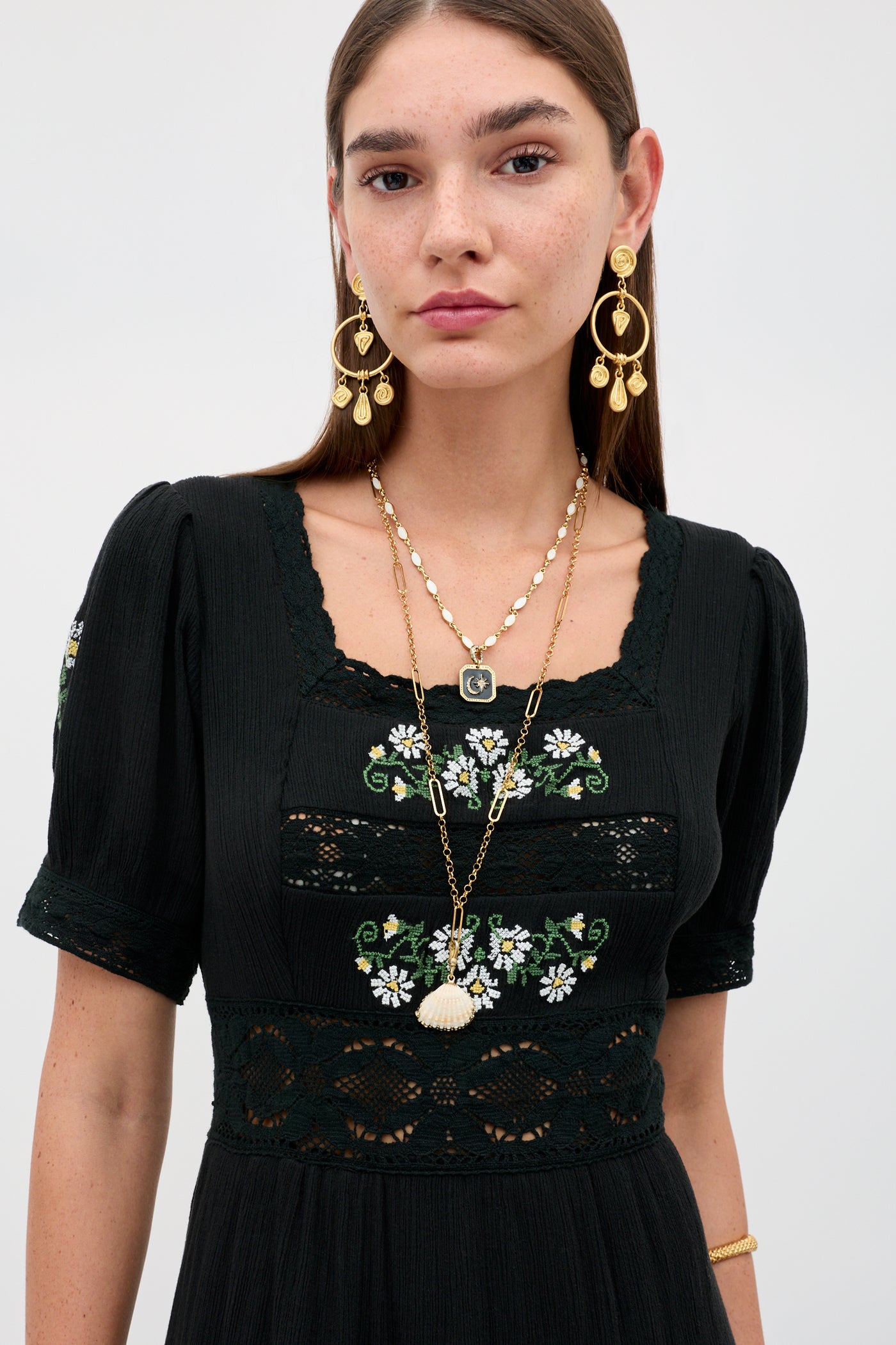 A woman with long brown hair wears the Philippa - Lilia Embroidery Black dress, featuring floral embroidery and lace details. She accessorizes with gold earrings, layered necklaces, and a bracelet while posing against a light backdrop.