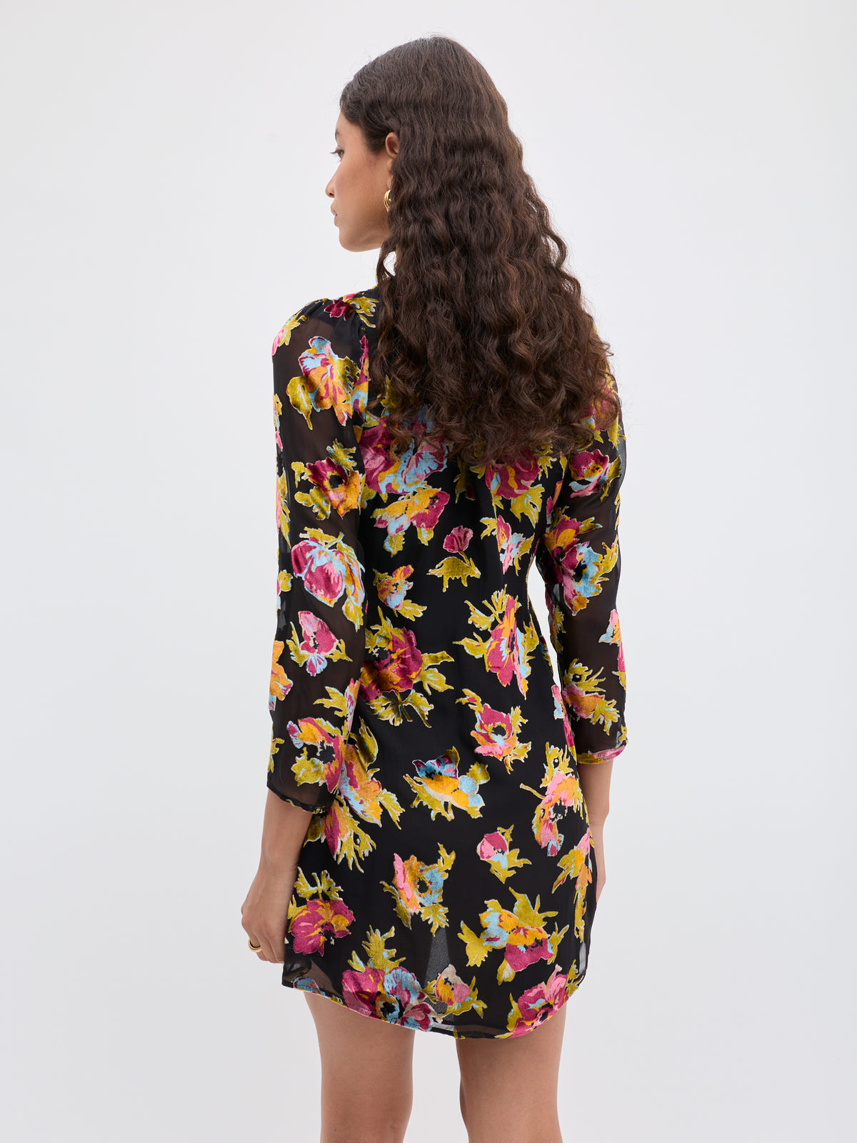 A woman with long, curly brown hair stands with her back to the camera, wearing the Kensley - Countryside Devoré Black dress—a black cocktail dress with colorful floral Devoré, long sleeves, and an above-the-knee hem against a plain white background.