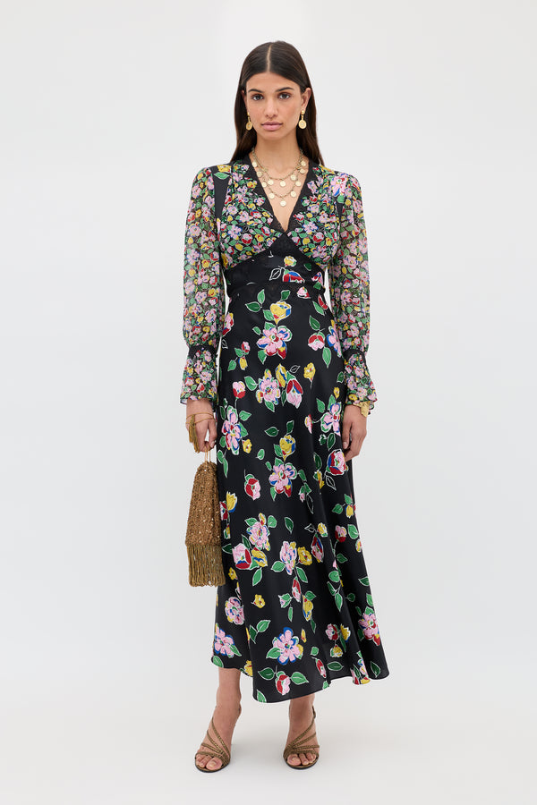 A woman stands against a white background wearing the Arlena - Kimmie Mix Black long-sleeved floral dress, nude heeled sandals, layered necklaces, hoop earrings, and holding a brown woven handbag.