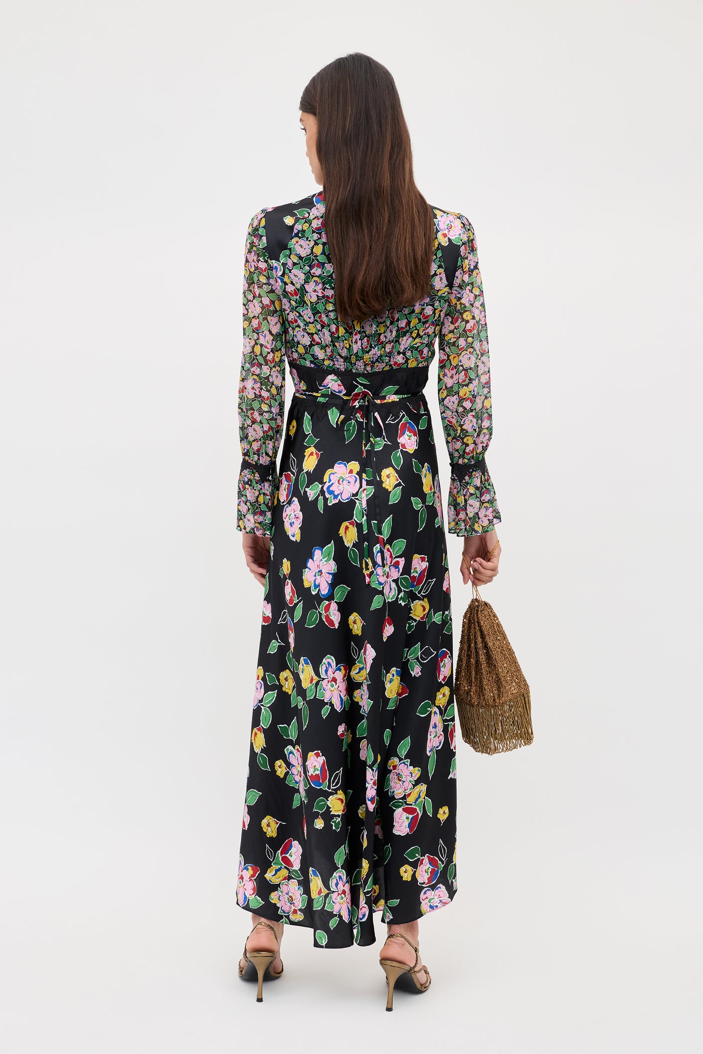 A woman with long brown hair faces away, wearing the Arlena - Kimmie Mix Black, a black maxi dress with colorful florals and long sleeves. She holds a woven handbag, wears black heeled sandals, and stands against a plain white background.