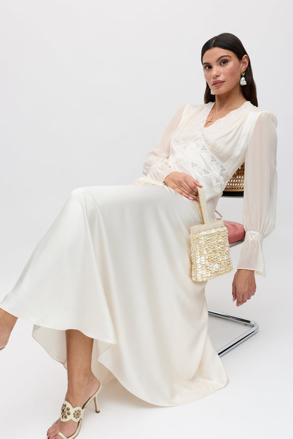 A woman with straight dark hair wears the Arlena - Pearl vintage midi dress with lace embroidery and sheer sleeves, paired with heeled sandals and statement earrings. She sits calmly on a pink chair, holding a woven handbag against a plain background.