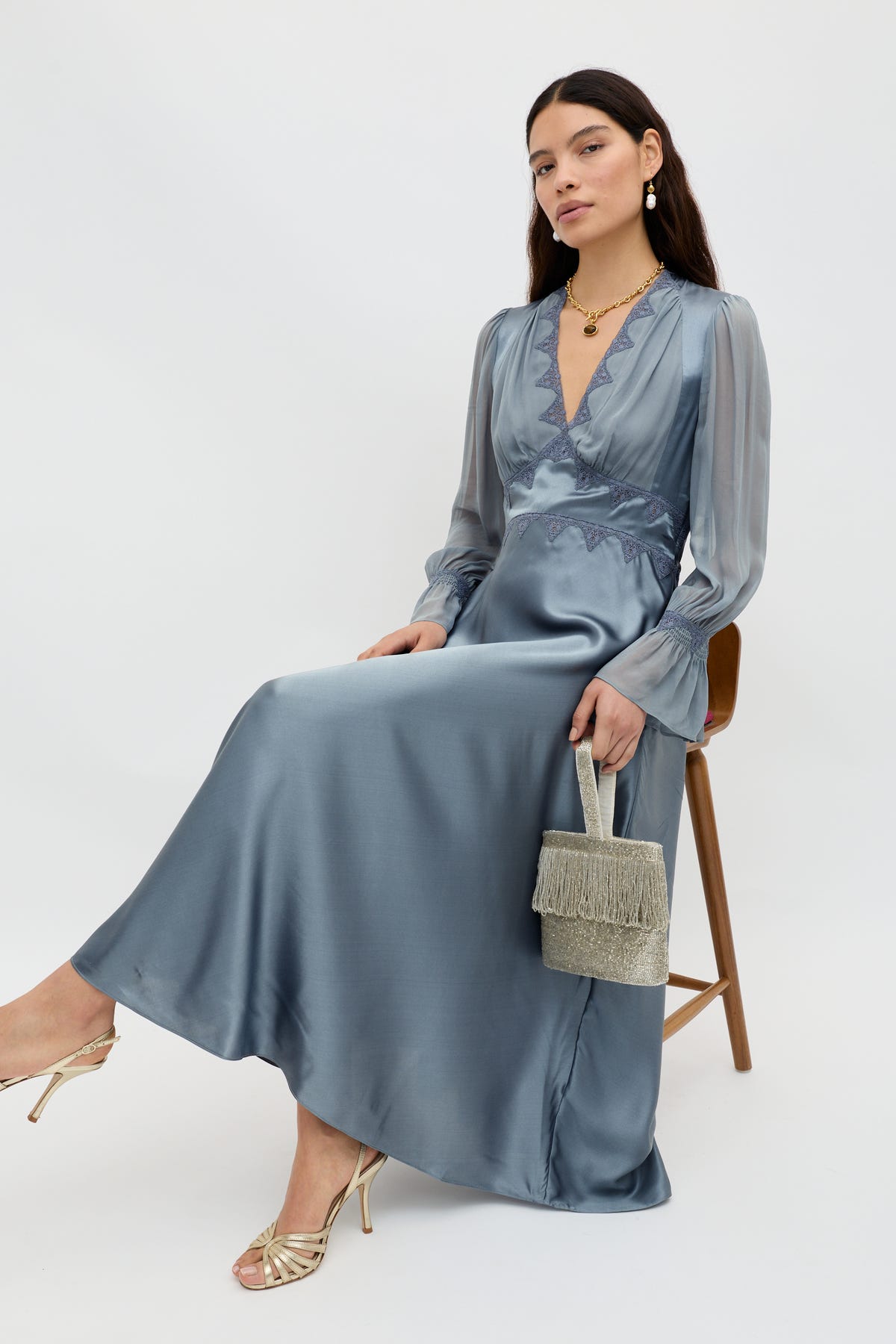 A woman sits on a wooden chair wearing the Arlena – Stone Blue silky, long-sleeved V-neck dress. She holds a fringed handbag and pairs the look with gold heels, a necklace, and earrings against a plain white background.