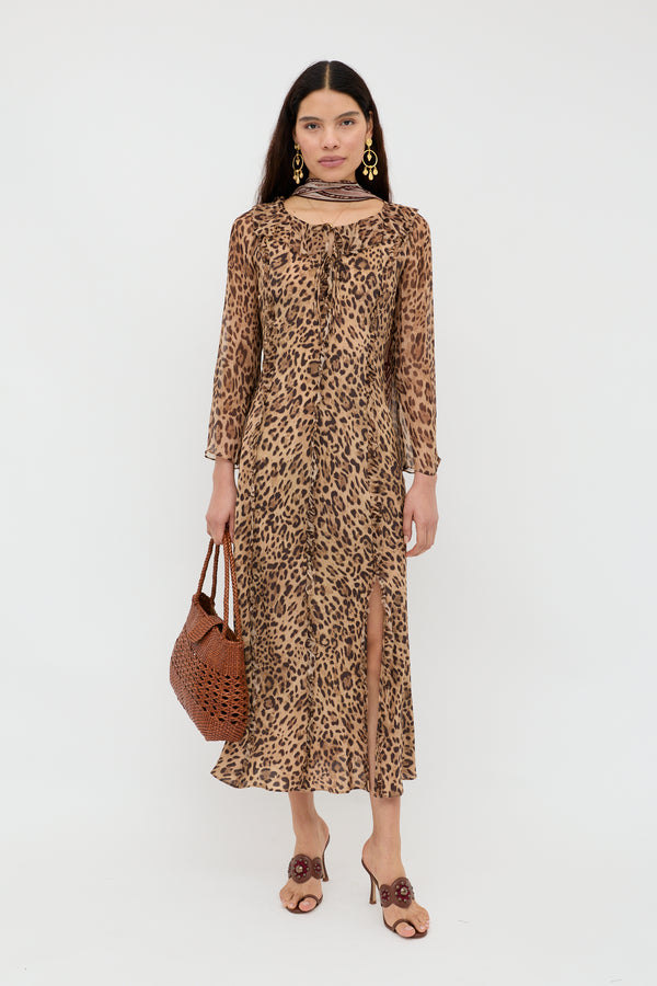 A woman stands against a white background wearing the Coralyn - Mila Leopard Brown 90s-inspired silk dress with long sleeves and a front slit, styled with large gold earrings, a brown woven bag, and beige heeled sandals with floral accents.