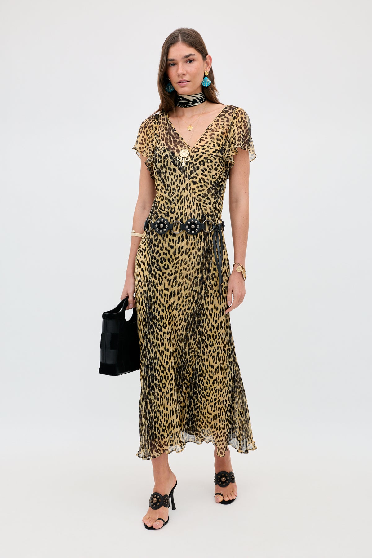 A woman poses against a plain background wearing the Bexley - Nina Leopard Brown dress, paired with black sandals, a black belt and handbag, turquoise earrings, a gold watch, layered necklaces, and a sheer black scarf.