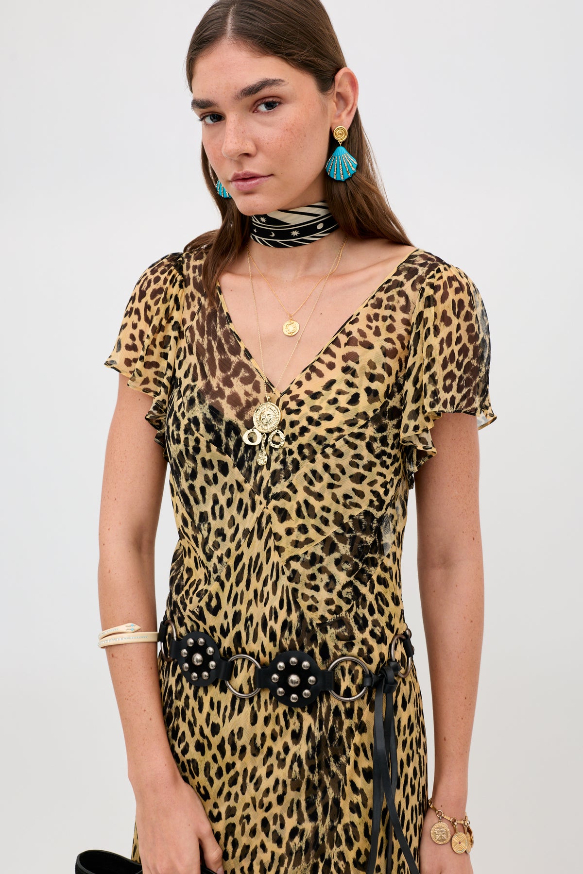 A woman wears the Bexley - Nina Leopard Brown dress with layered gold necklaces, blue tassel earrings, a black choker, a studded black belt, and holds a black bag at her side against a plain background.