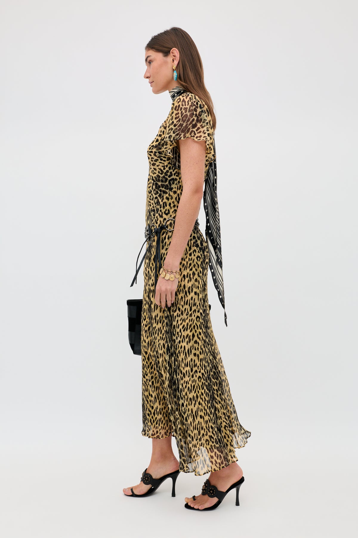 A woman in profile wears the Bexley - Nina Leopard Brown dress, black heeled sandals, turquoise earrings, a black handbag, and a neck scarf against a plain white background.