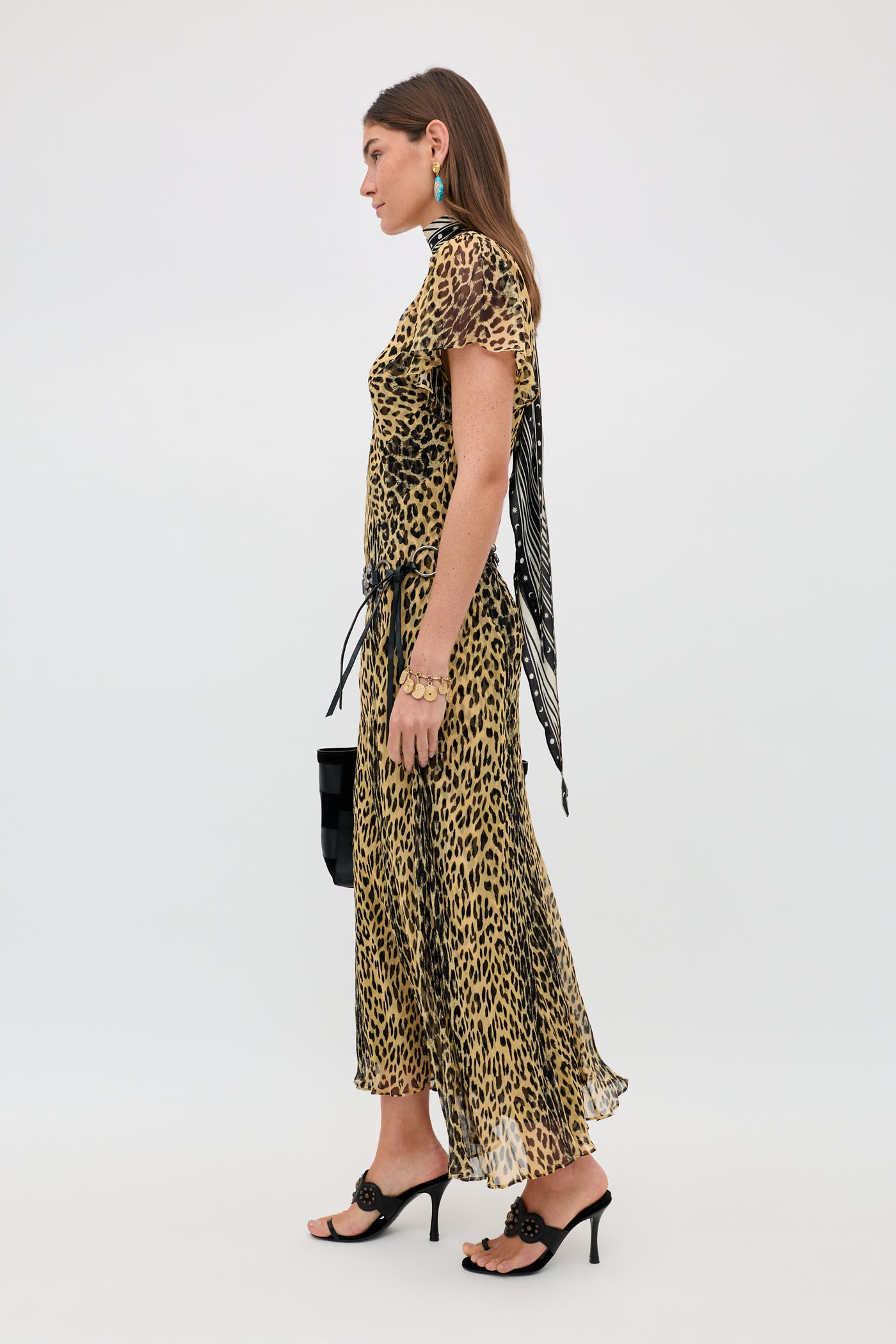 A woman in profile wears the Bexley - Nina Leopard Brown dress, black heeled sandals, turquoise earrings, a black handbag, and a neck scarf against a plain white background.