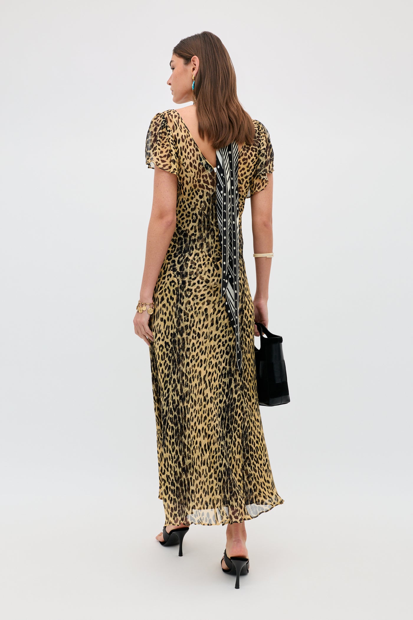 A woman faces away, wearing the Bexley - Nina Leopard Brown dress with a low back and black-and-white tie, paired with a black handbag, black heels, gold jewelry, and drop earrings. The background is plain and light-colored.