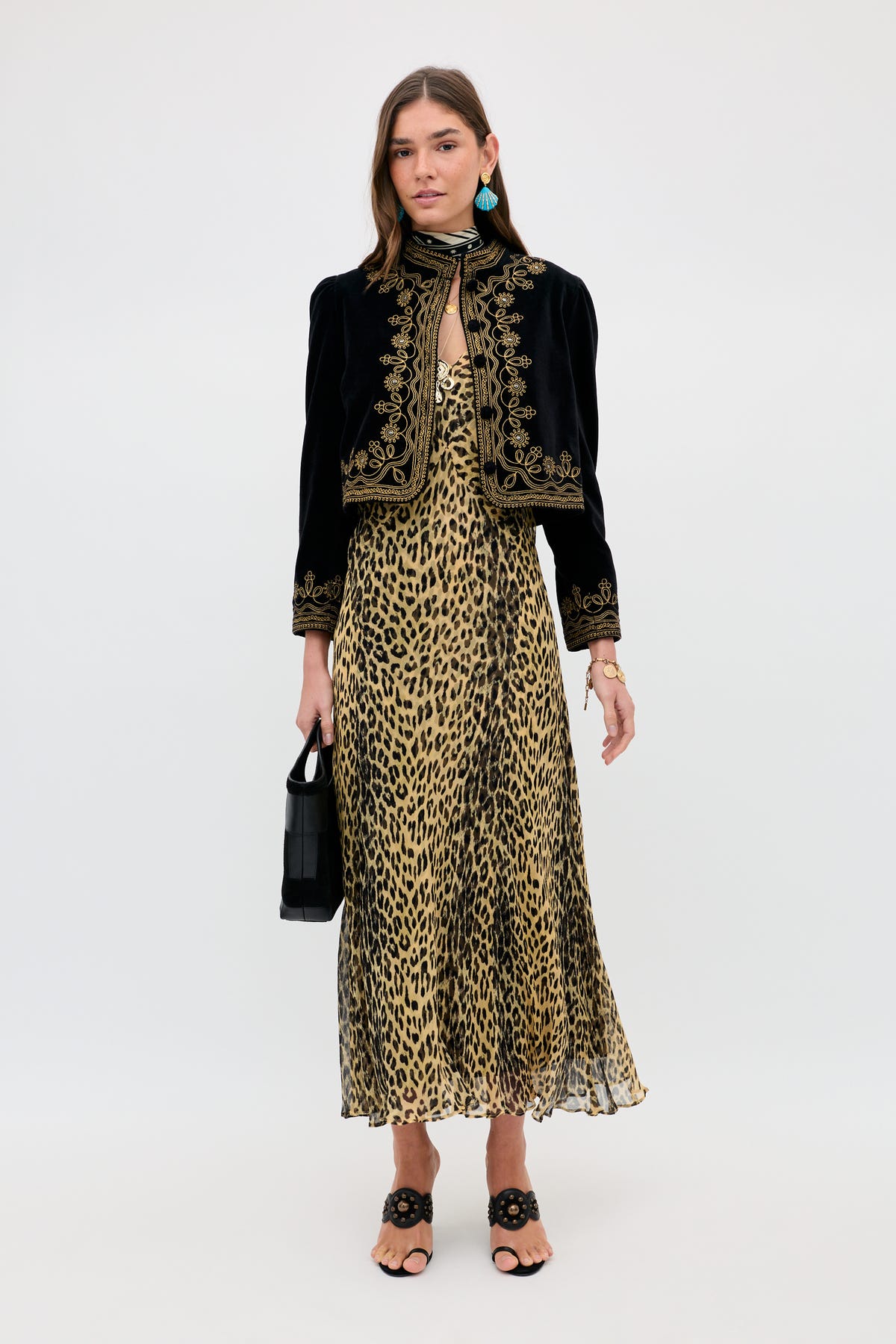 A woman with shoulder-length brown hair wears the Bexley - Nina Leopard Brown dress, a black embroidered jacket, blue earrings, black open-toe sandals, and holds a black handbag. She stands against a plain white background.