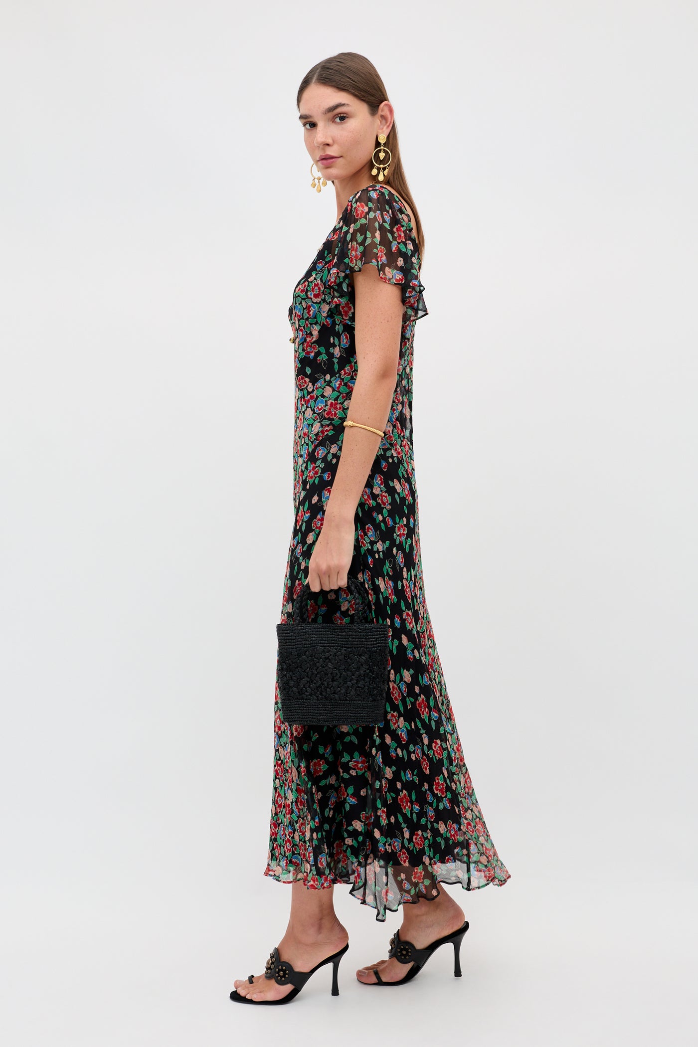 A woman in the Bexley - Jeannie Mix Charcoal black, ankle-length floral dress with short flutter sleeves, black heeled sandals, gold jewelry, and a textured black handbag stands against a plain white background.