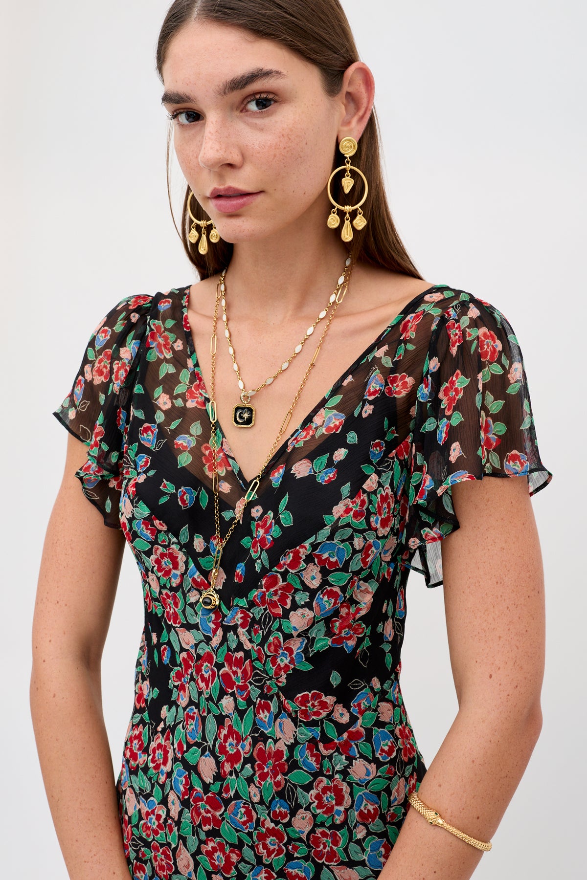 A woman with long brown hair wears the Bexley - Jeannie Mix Charcoal dress, featuring red, green, and blue floral patterns, paired with layered gold necklaces, large gold earrings, and a gold bracelet against a plain background.