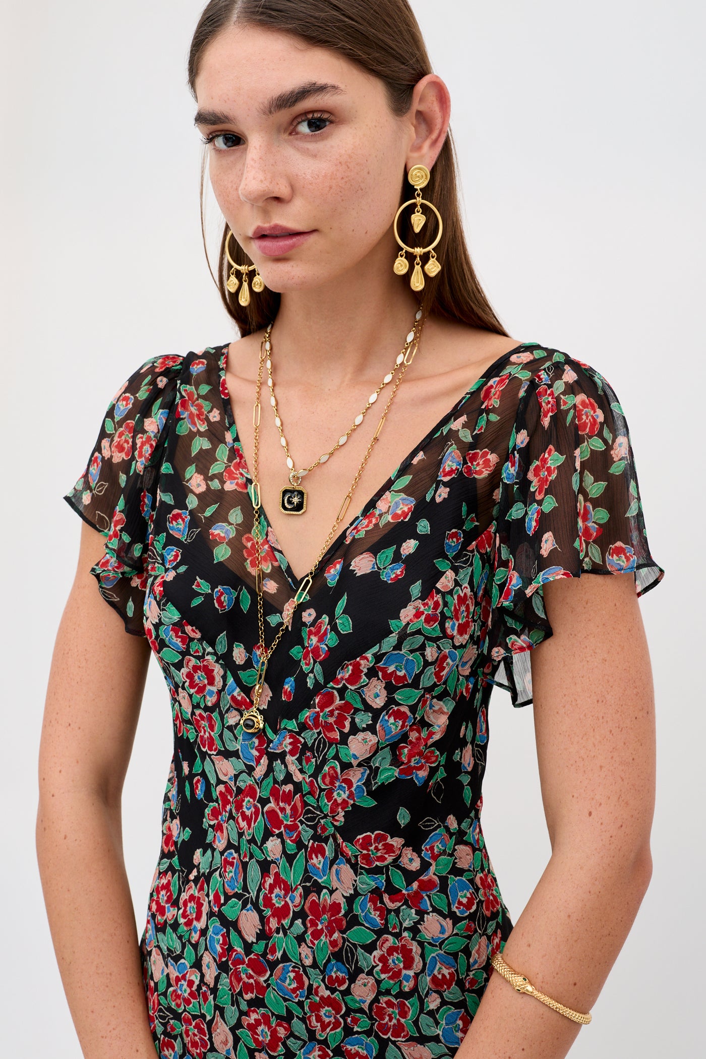 A woman with long brown hair wears the Bexley - Jeannie Mix Charcoal dress, featuring red, green, and blue floral patterns, paired with layered gold necklaces, large gold earrings, and a gold bracelet against a plain background.