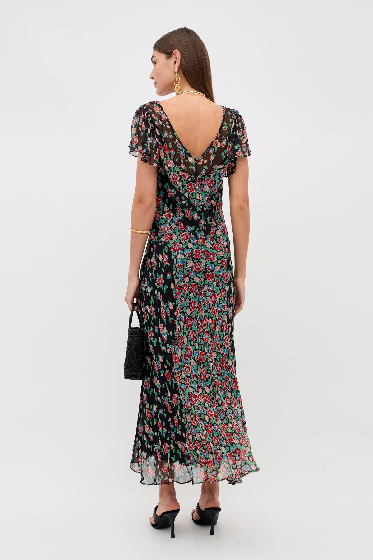 A woman faces away, wearing the Bexley - Jeannie Mix Charcoal dress featuring a V-back, flutter sleeves, and flowing skirt. She carries a black handbag, wears gold jewelry and black heels. The background is plain and light.