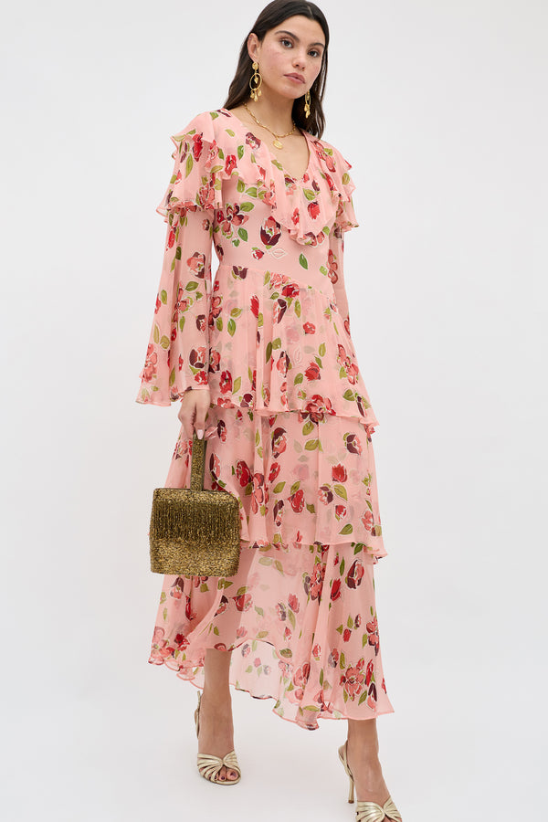A woman wearing the Loralyn - Kimmie Floral Pink dress with gold earrings, gold open-toe heels, and a small gold textured handbag stands against a plain white background.