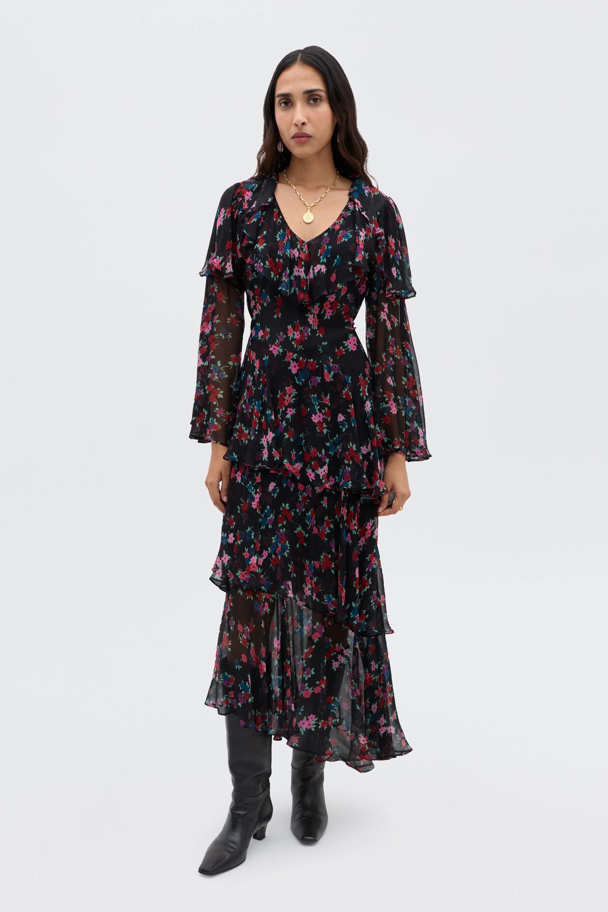 A woman wearing the Loralyn - City Floral Black long, boho dress with layered ruffles and sheer sleeves, paired with black knee-high boots, stands against a plain white background.