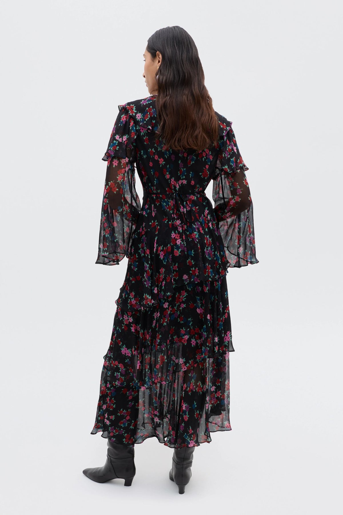 A person with long dark hair faces away, wearing the Loralyn - City Floral Black dress with a colorful floral pattern and ruffled sleeves, paired with black boots against a plain light background.