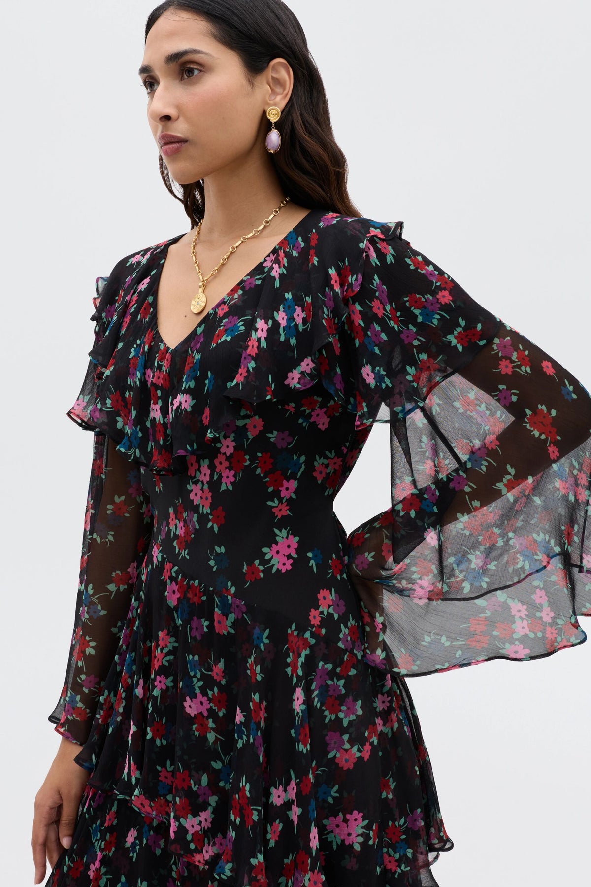 A woman wears the Loralyn - City Floral Black dress, a sheer black boho style featuring colorful florals, ruffled details, and a V-neck. She completes her look with gold earrings and a necklace against a plain background.
