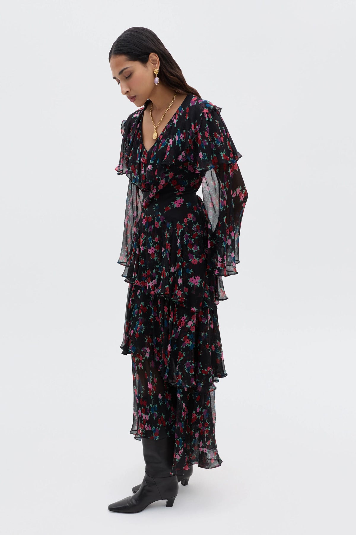 A woman stands sideways wearing the Loralyn - City Floral Black dress with layered sleeves and skirt, paired with black boots. She looks down with her hands by her sides against a plain light background.