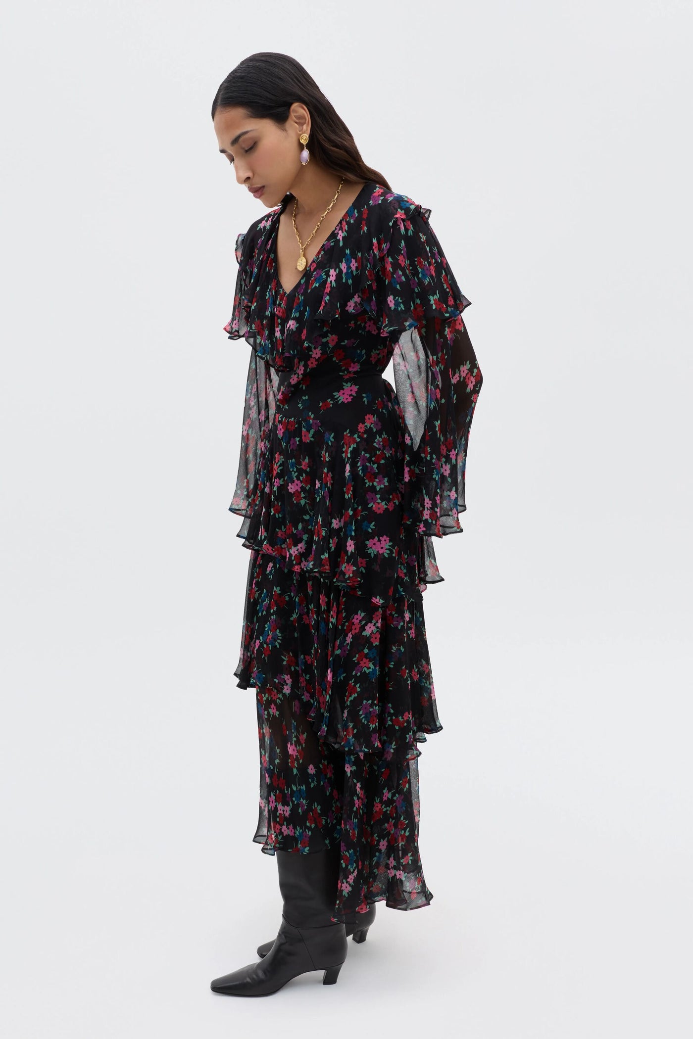 A woman stands sideways wearing the Loralyn - City Floral Black dress with layered sleeves and skirt, paired with black boots. She looks down with her hands by her sides against a plain light background.