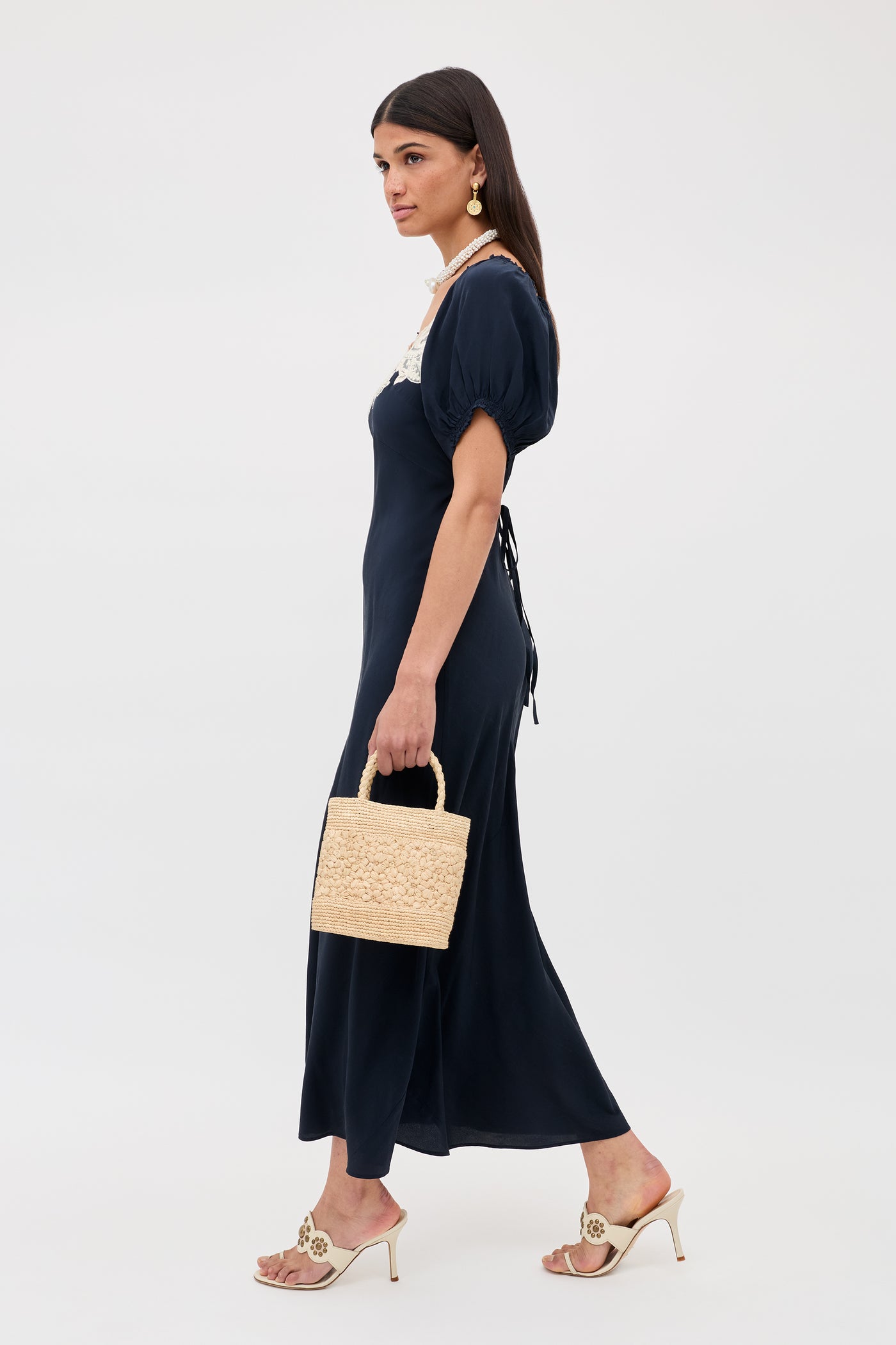 A woman wears the Ilaria - Deep Navy dress, featuring short sleeves and ankle length, paired with a small woven handbag, cream heeled sandals, and statement earrings. Her dark hair is down against a plain white background.