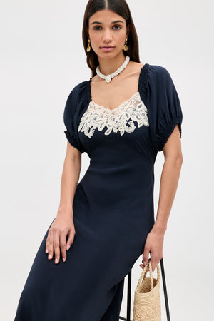 A woman in the Ilaria - Deep Navy dress with puffed sleeves and white lace neckline detail, accessorized with pearls and a woven straw bag, sits on a stool against a plain background.
