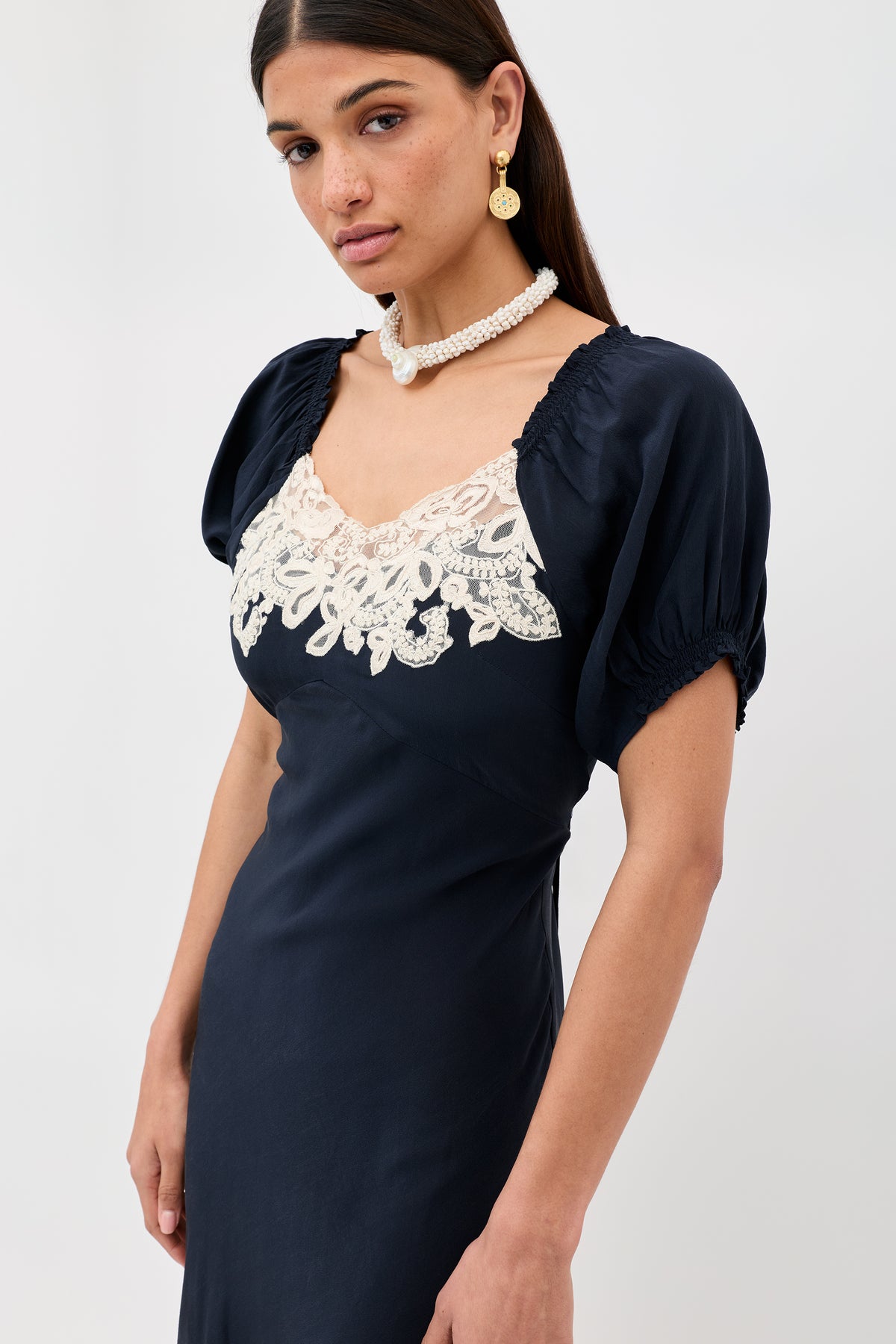 A woman wearing the Ilaria - Deep Navy dress with short puff sleeves and white lace bodice detail, accessorized with pearl earrings and a matching necklace, stands against a plain white background.
