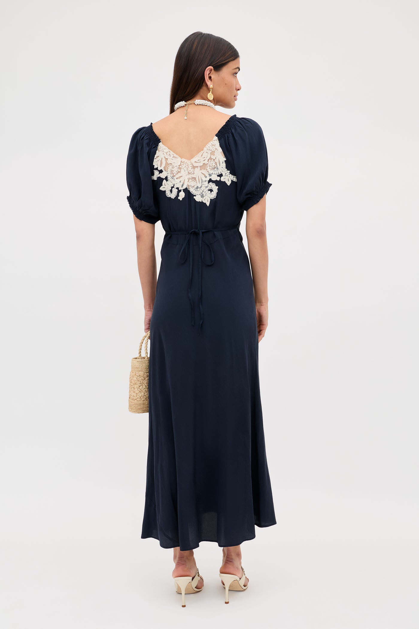 A woman stands facing away, wearing the Ilaria - Deep Navy dress—an ankle-length style with short sleeves, a V-shaped back, and white lace accents. She carries a straw handbag, wears beige heels, and accessorizes with a pearl necklace.
