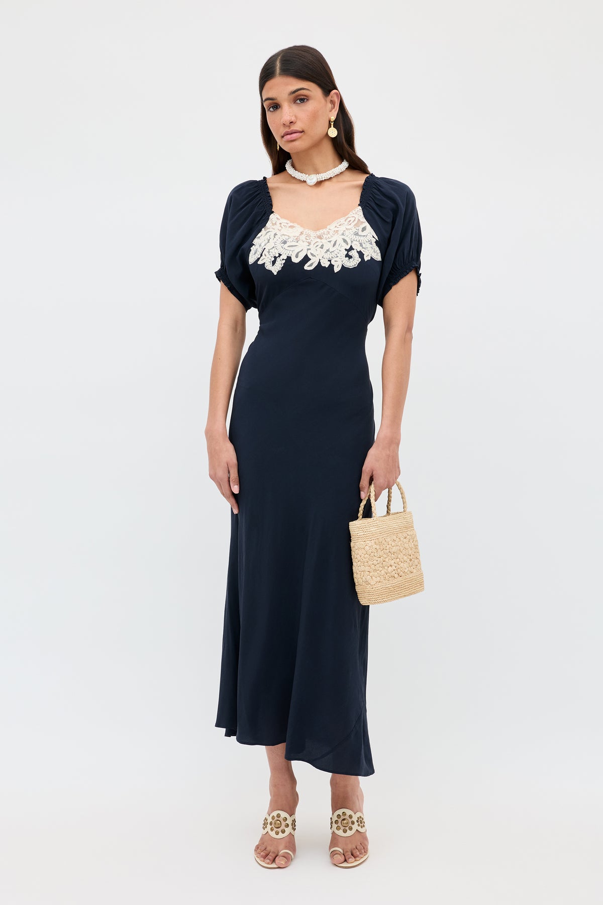 A woman wearing the Ilaria - Deep Navy midi dress with white lace neckline, light heeled sandals, a straw handbag, and a shell necklace stands against a white background.