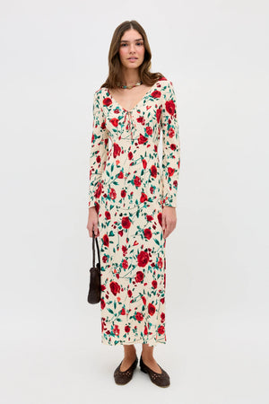 A woman stands against a plain white background wearing the Gwenyth - Claudia Rose Cream dress with red rose and green leaf print, brown flats, a necklace, and holding a small brown handbag.