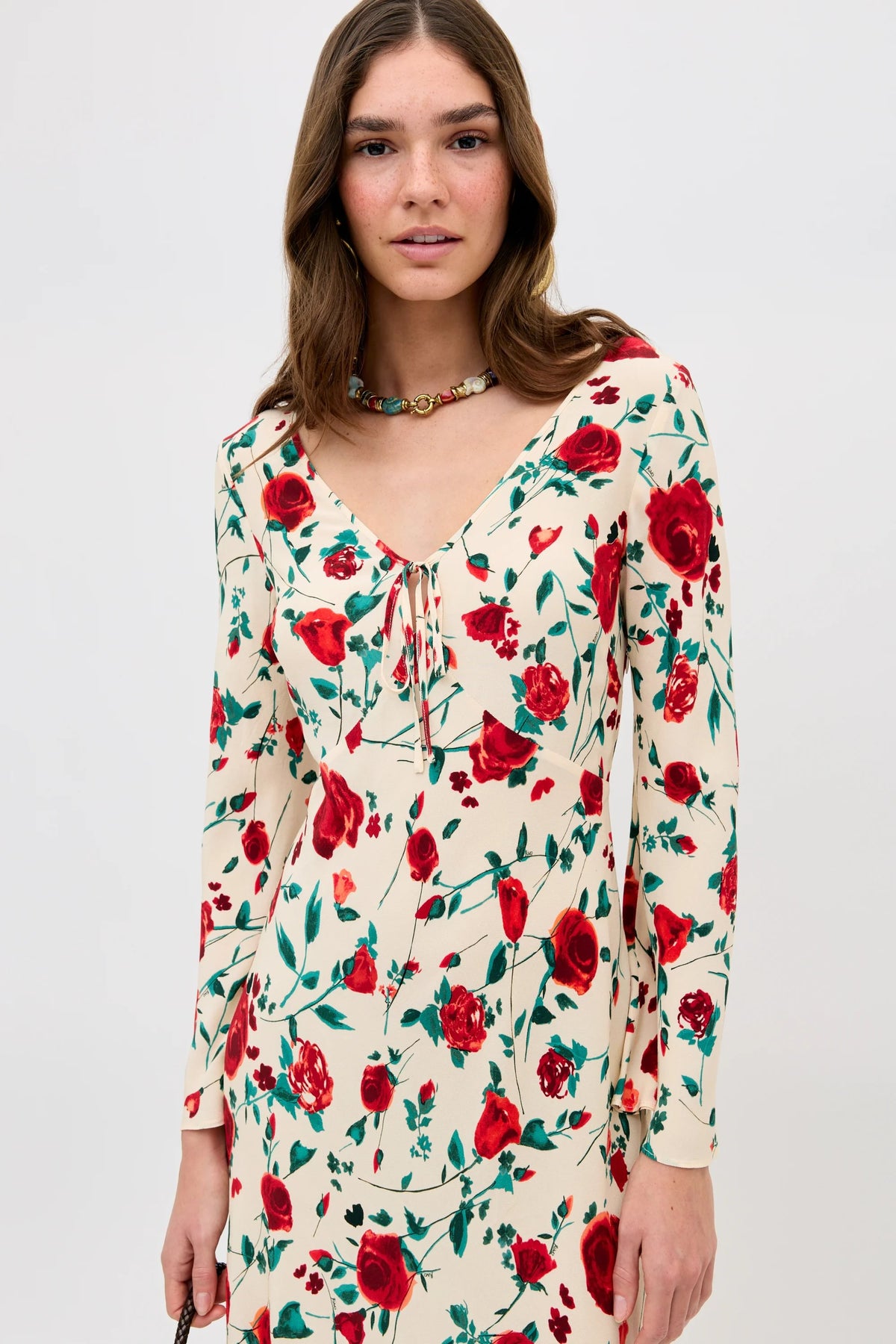 A woman with long brown hair wears the Gwenyth - Claudia Rose Cream dress, featuring red rose and green leaf prints with a small neckline bow, and a colorful beaded necklace against a plain white background.