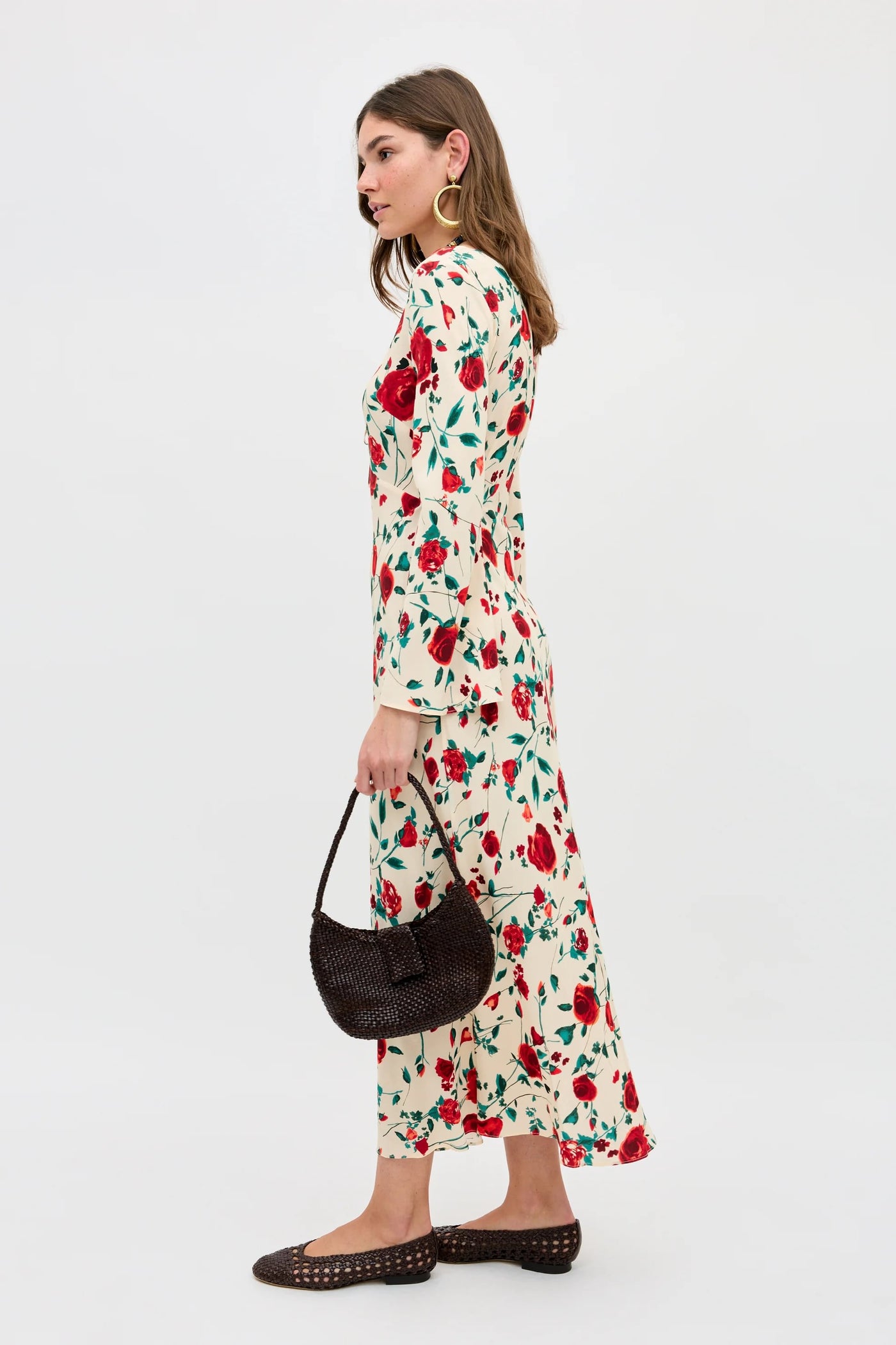 A woman stands sideways against a white background wearing the Gwenyth - Claudia Rose Cream, a long cream dress with red and green floral pattern. She accessorizes with a dark woven handbag, brown flats, large hoop earrings, and has shoulder-length hair.