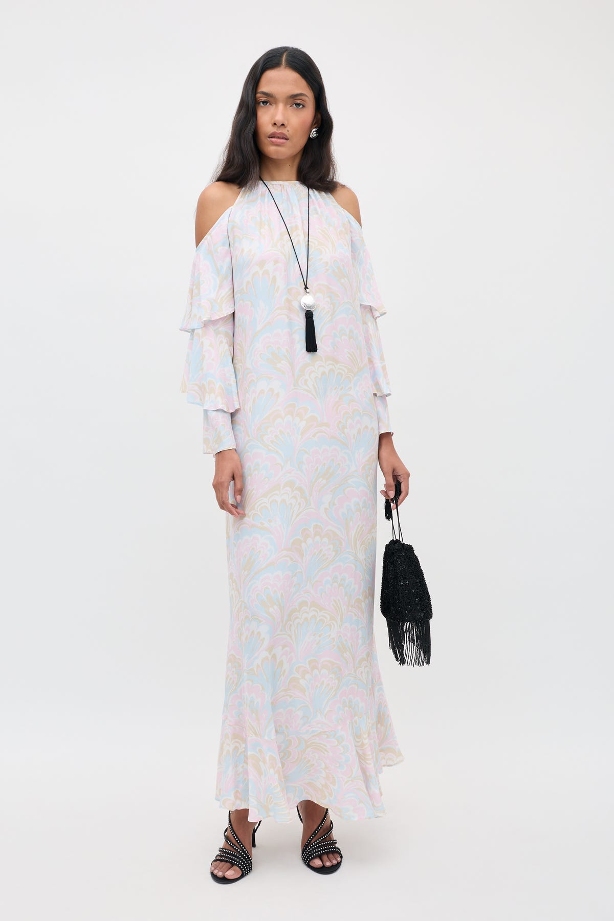 A woman in the Niara - Marble Swirl Pastel cold-shoulder maxi dress with ruffled sleeves, holding a black fringed bag, wearing black sandals and a long necklace, stands against a plain white background.