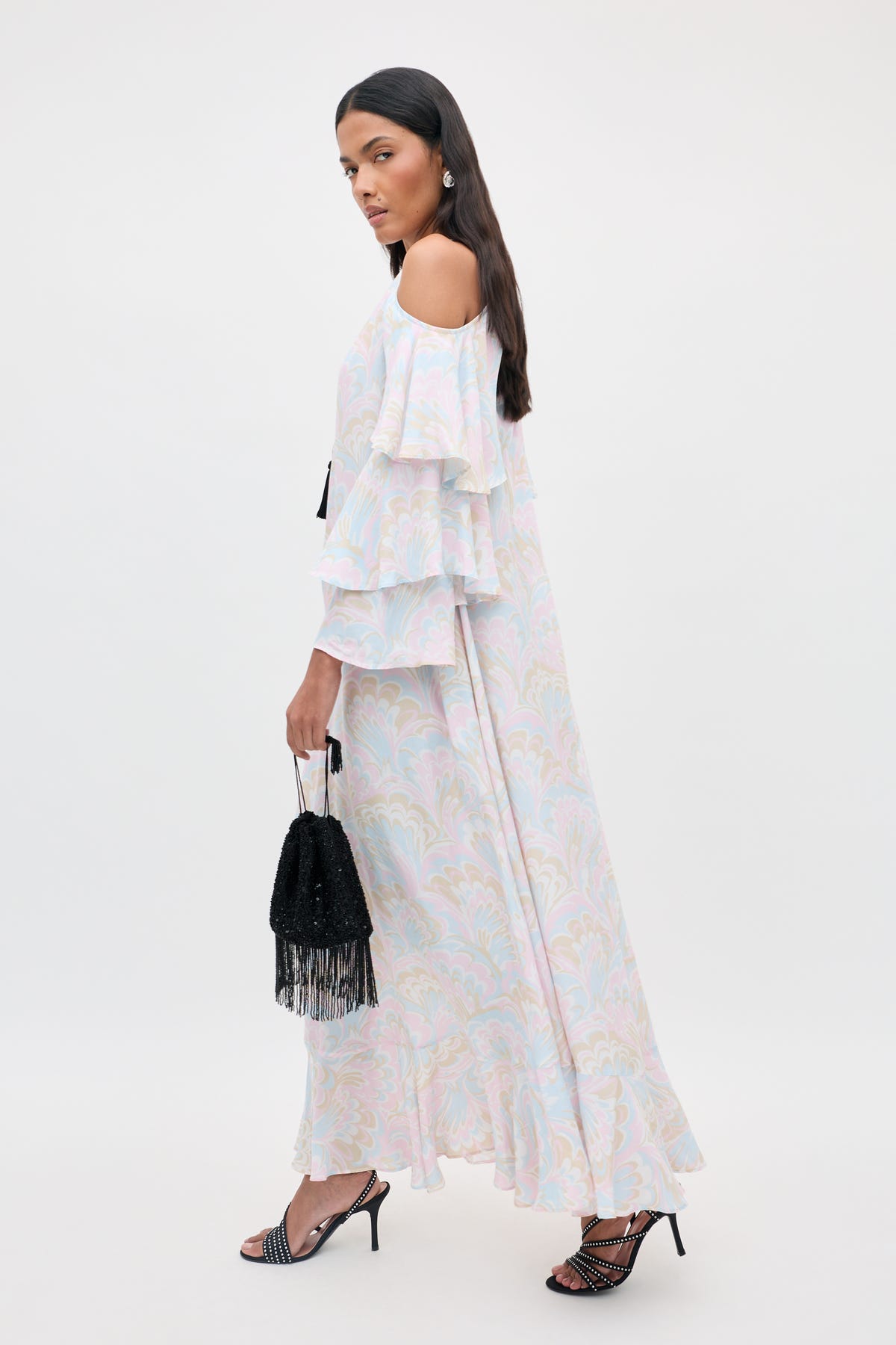 A woman stands against a plain light background wearing the Niara - Marble Swirl Pastel dress: a long, off-shoulder design with ruffled sleeves in a soft pastel marble pattern. She accessorizes with a black beaded fringe bag and black strappy heels.