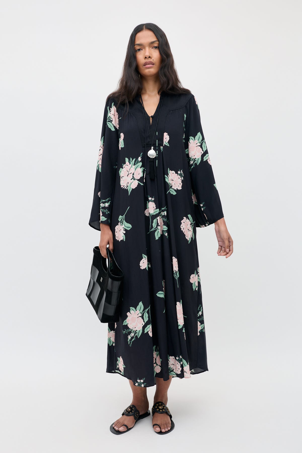 A woman wearing the Lucilla - Peonies Scatter Black dress, featuring pink and green floral patterns, paired with black sandals and a black handbag, stands against a plain light background.