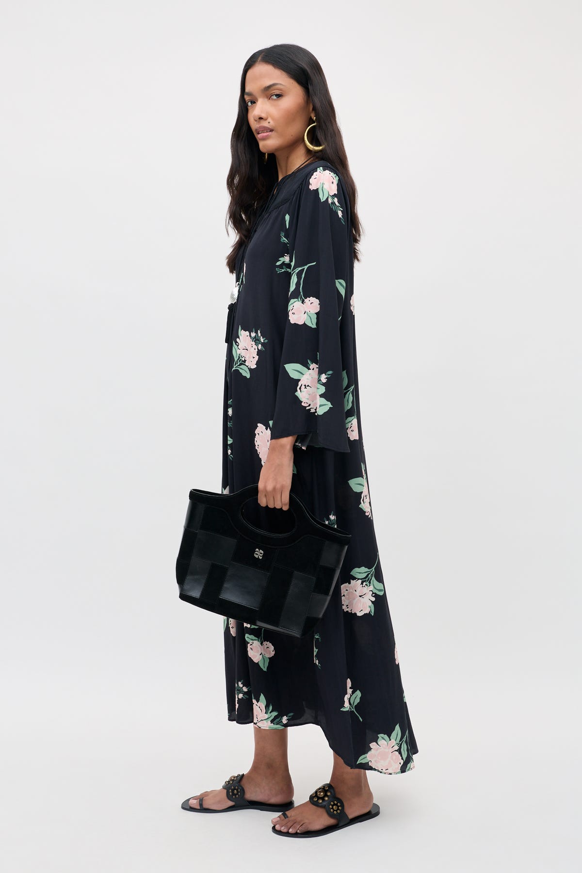 A woman wearing the Lucilla - Peonies Scatter Black dress with loose sleeves, paired with a black handbag, black sandals, and large hoop earrings, stands against a plain light background.