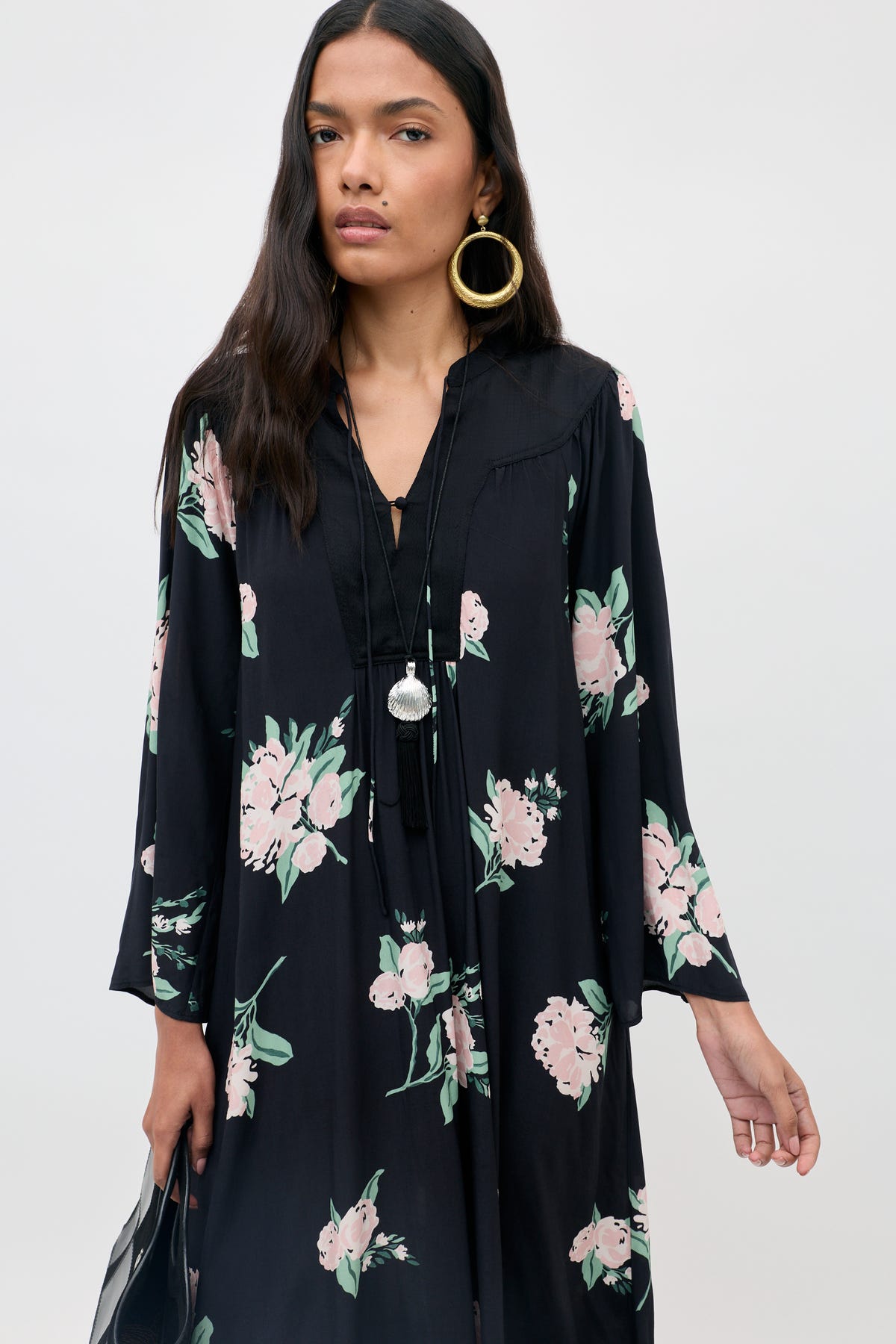 A woman wearing the Lucilla - Peonies Scatter Black dress, featuring black fabric with pink floral patterns, styled with large hoop earrings and a long necklace, stands against a plain background.