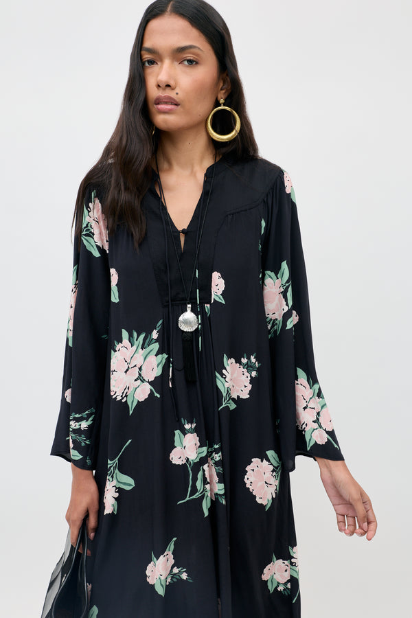 A woman wearing the Lucilla - Peonies Scatter Black dress, featuring black fabric with pink floral patterns, styled with large hoop earrings and a long necklace, stands against a plain background.