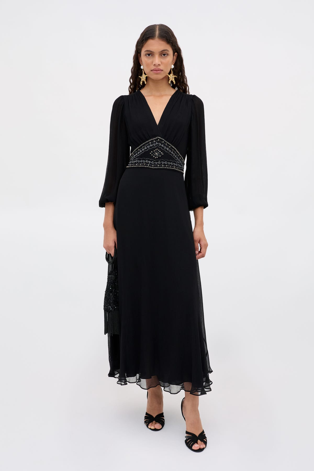A woman stands against a white background, wearing the Kaesha - Diana Embellishment Black long v-neck dress with three-quarter sleeves and a beaded waistband, paired with black open-toe heels, and holding a black bag in her left hand.