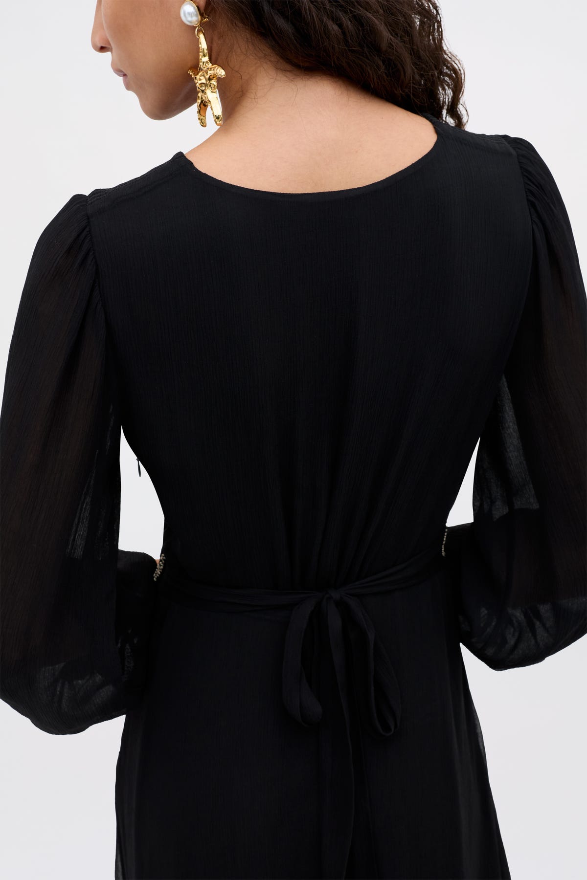 A woman with long, wavy brown hair models the Kaesha - Diana Embellishment Black dress, featuring sheer long sleeves and a tie waist. She accessorizes with large gold statement earrings adorned with pearls.