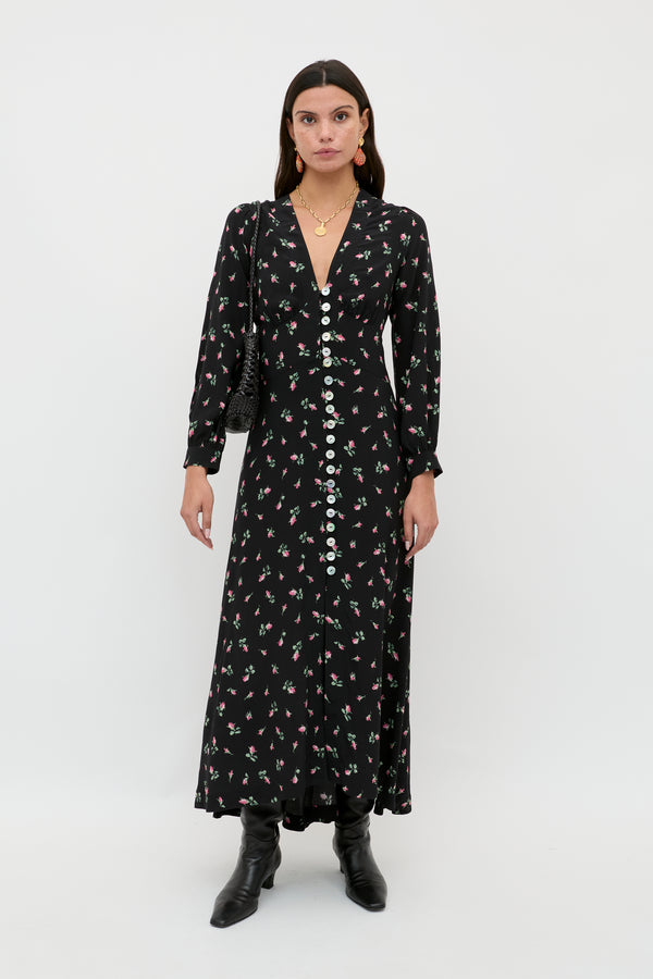 A woman wears the Avalyn - Sienna Bud Black long sleeve midi dress with vintage-inspired cuffs, paired with black boots, a small black purse, and gold jewelry, standing against a plain white background.