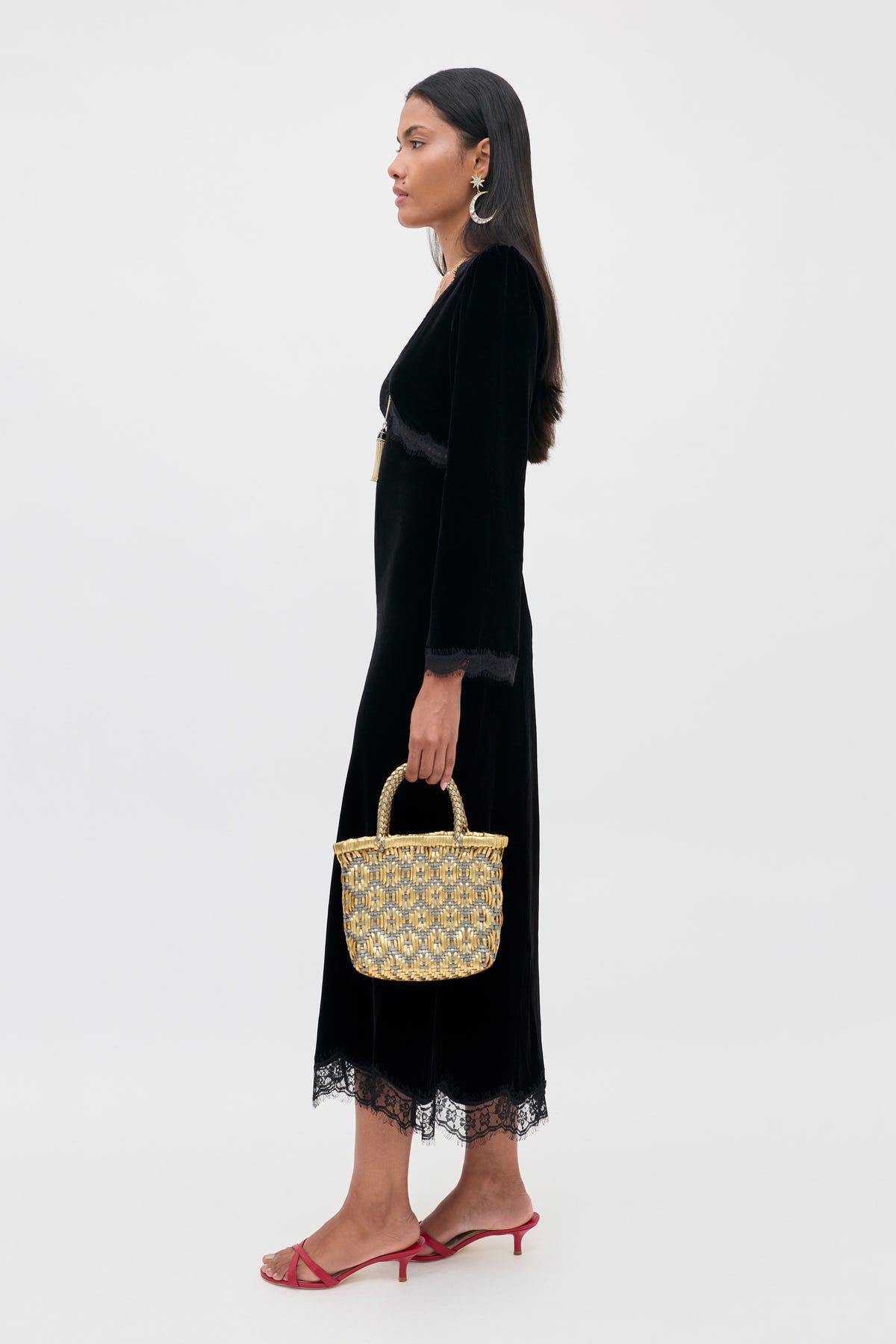 A woman in profile wears an Alessi - Black long-sleeved velvet midi dress with lace detailing and a boho-glamour aesthetic, paired with a small metallic gold woven basket bag and red heeled sandals.