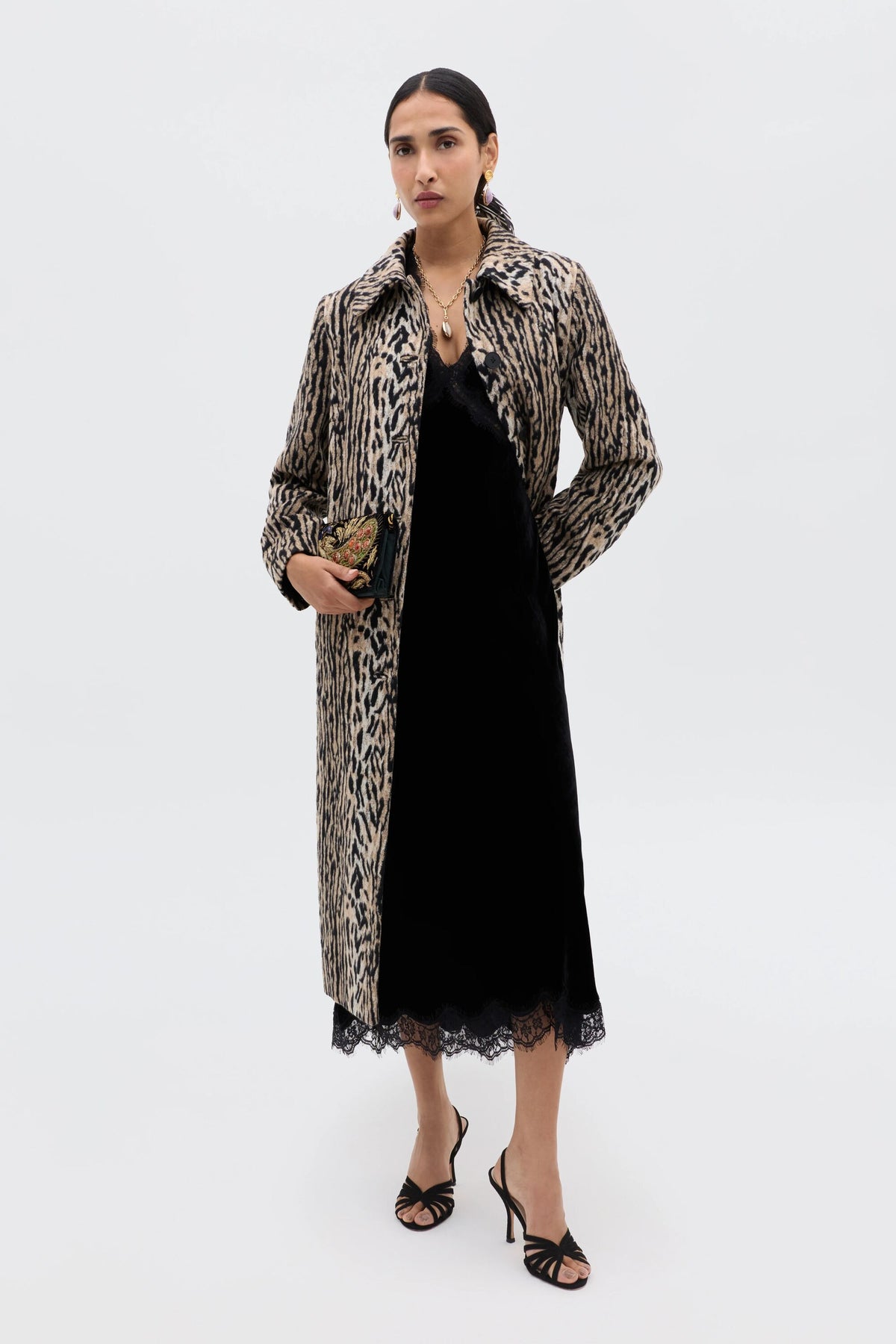 A woman poses against a white background in the Alessi - Black velvet dress with lace detailing, layered under a long leopard-print coat. She holds a small clutch, wears black strappy heels, and has her dark hair tied back.