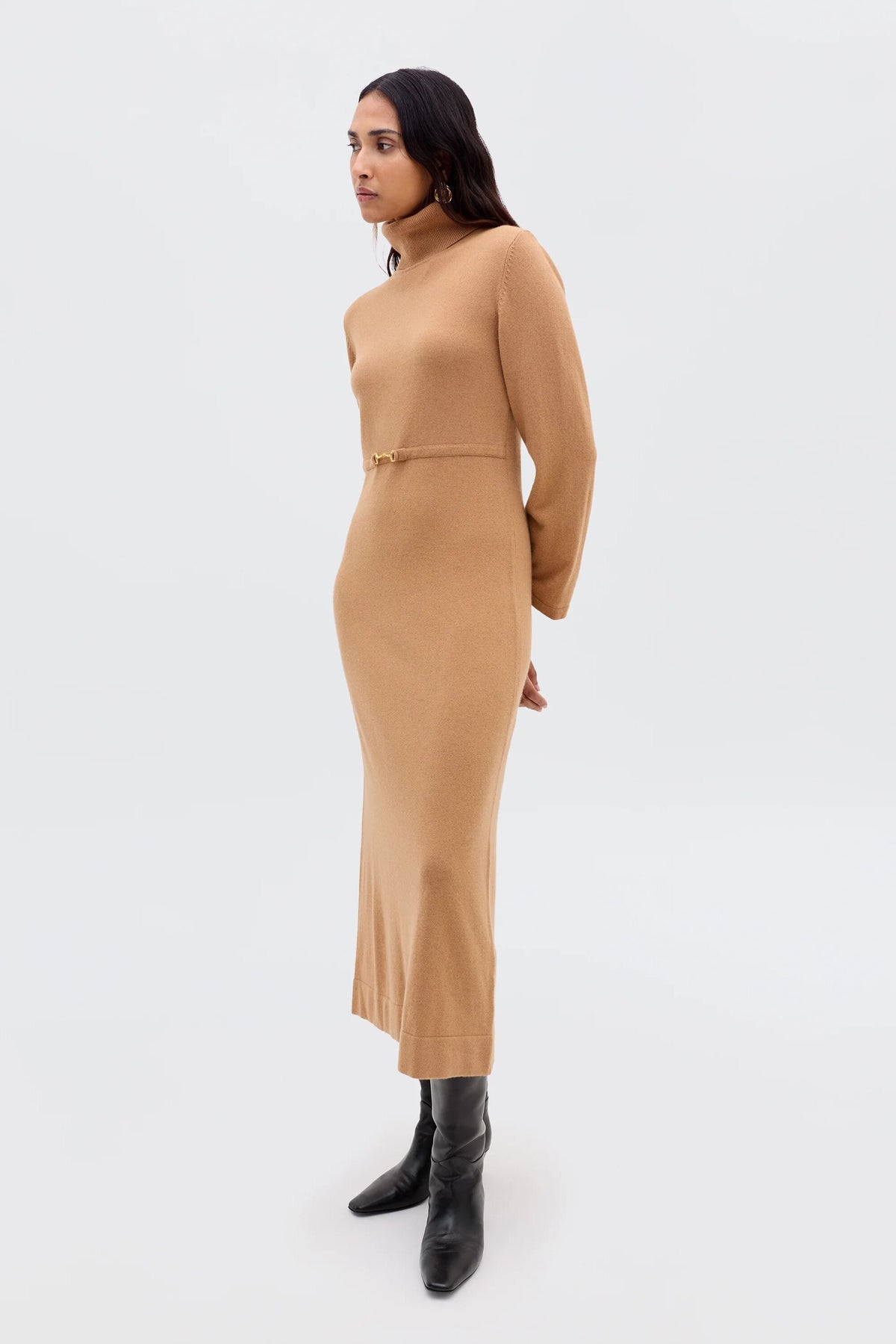 A woman stands against a plain background wearing Elliette - Camel, a minimalist camel-colored knitted dress with long sleeves and a fitted waist, styled with black knee-high boots. She faces slightly to the side, arms relaxed by her sides.