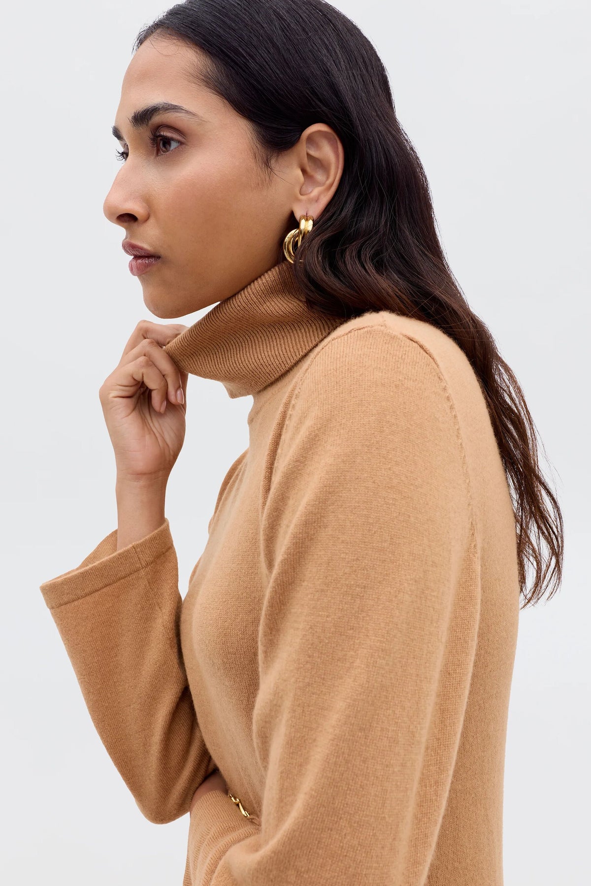 A woman with long dark hair and gold hoop earrings wears the Elliette - Camel turtleneck dress, holding the collar as she poses in profile against a light, neutral background, reflecting a minimalist style.