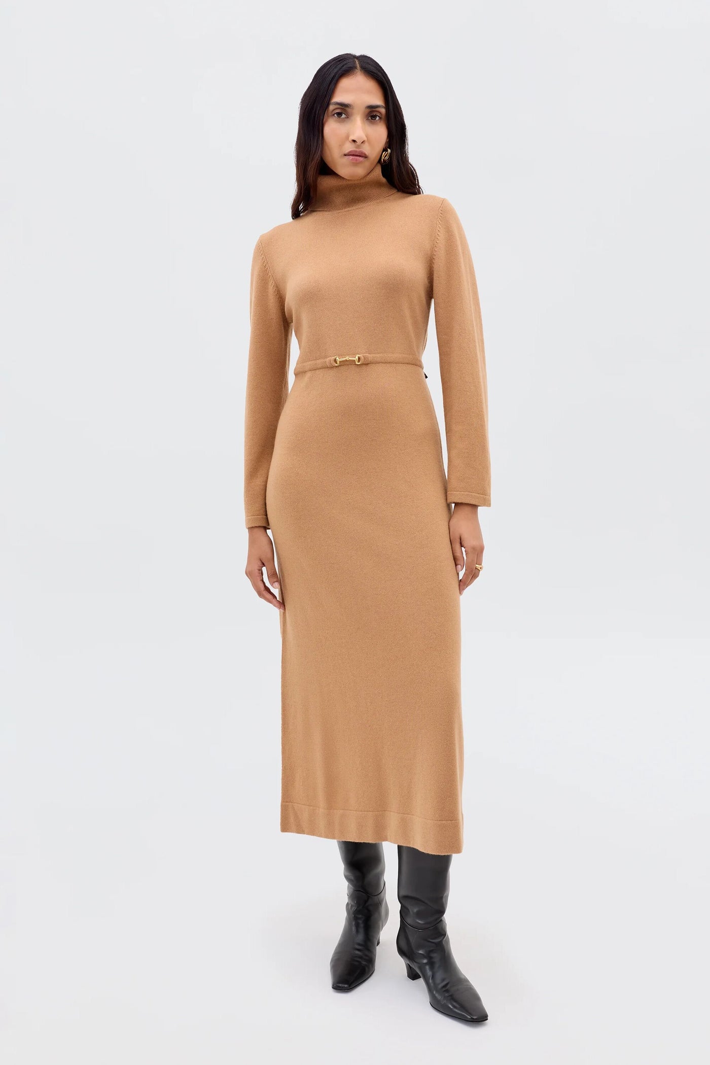 A woman wears the Elliette - Camel, a long-sleeved knitted dress with a slim waist belt, paired with black knee-high boots, standing against a white background to highlight its timeless minimalist style.