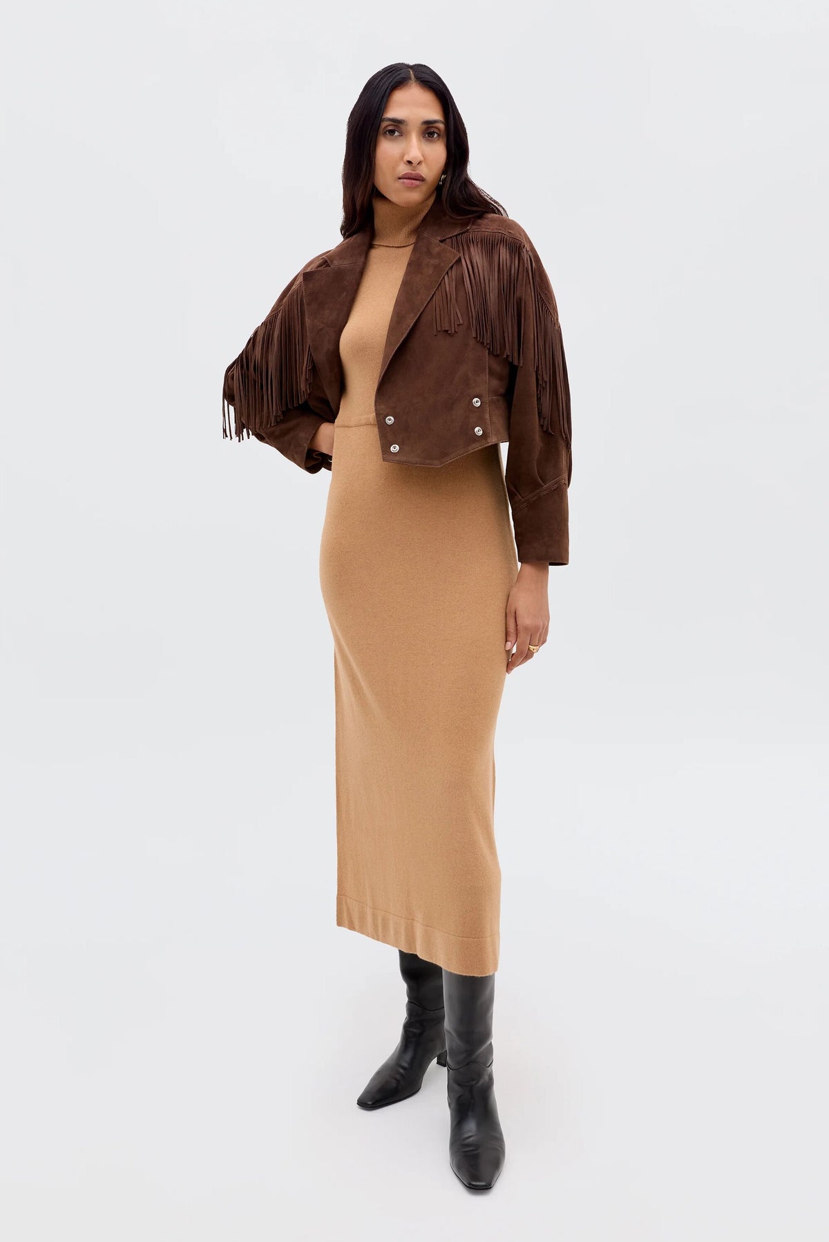 A woman stands against a plain background wearing the Elliette - Camel, a long, fitted tan turtleneck dress. She pairs it with a brown suede fringe jacket and knee-high black boots, embodying effortless minimalist style.