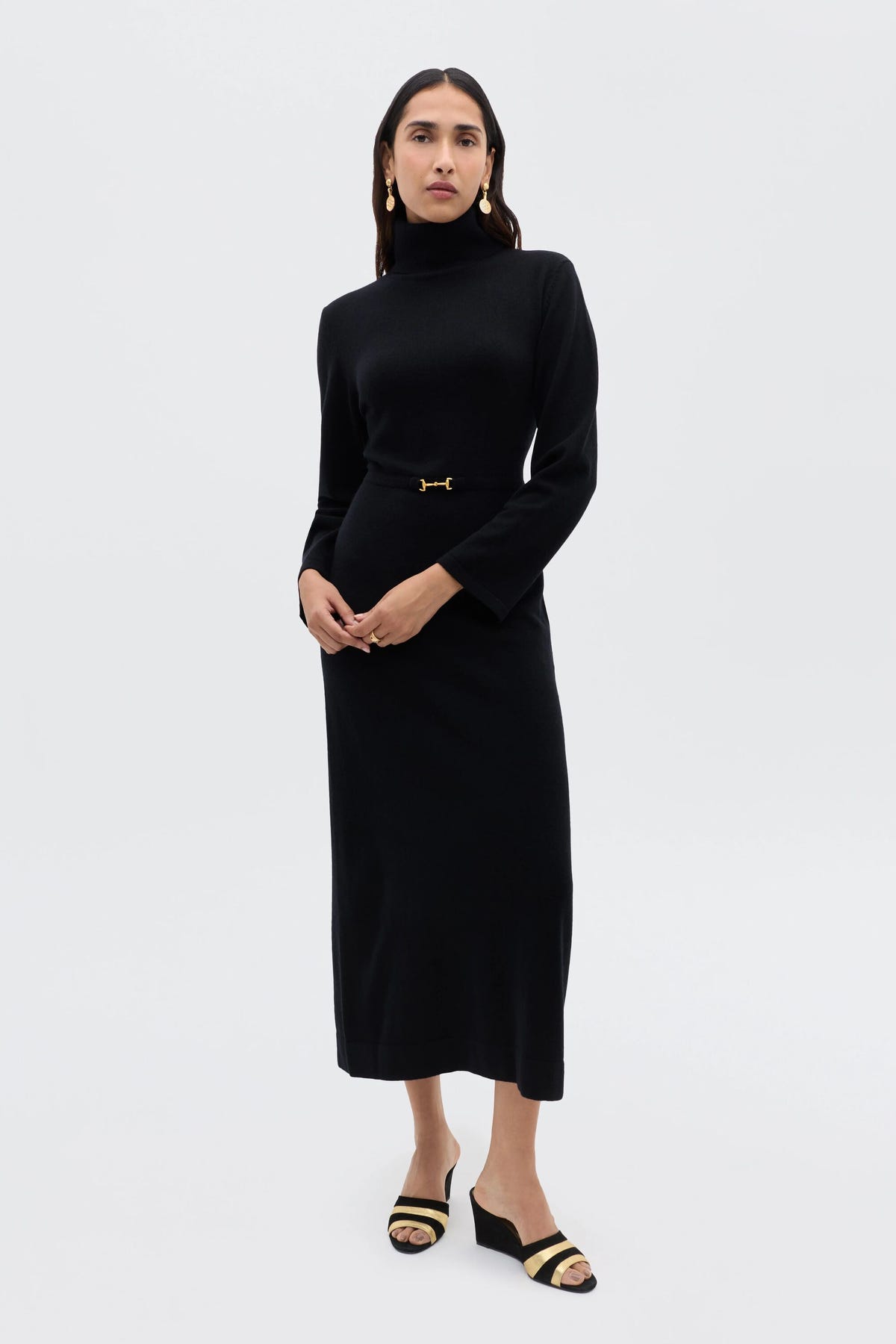 A woman with straight dark hair and a neutral expression poses against a light background, wearing the Elliette - Black long-sleeved turtleneck dress with gold waist detail, styled with black open-toe sandals featuring gold accents.