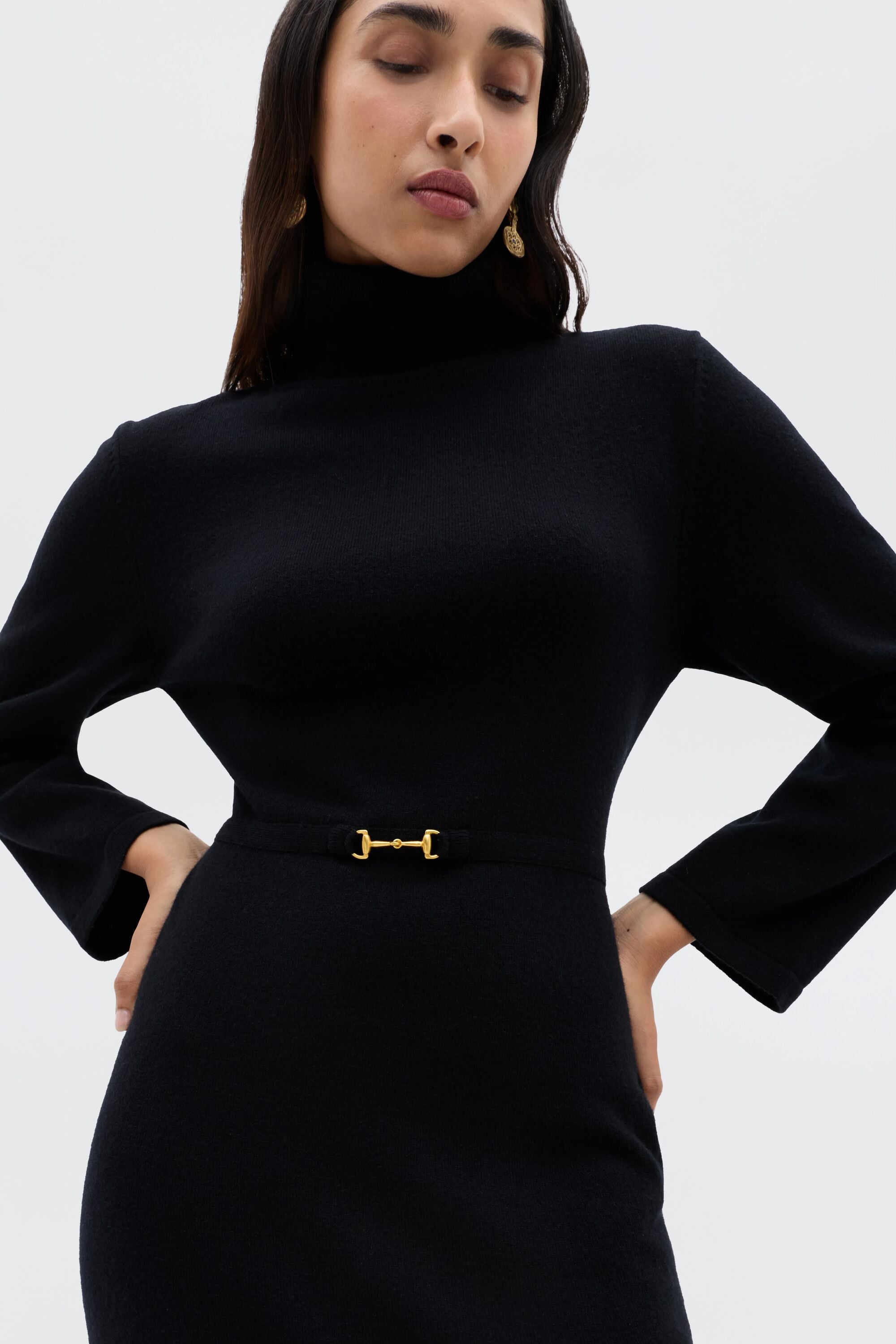 A woman models the Elliette - Black, a fitted black knitted turtleneck dress with long sleeves and a gold waist buckle, highlighting its refined minimalist style against a plain white background.